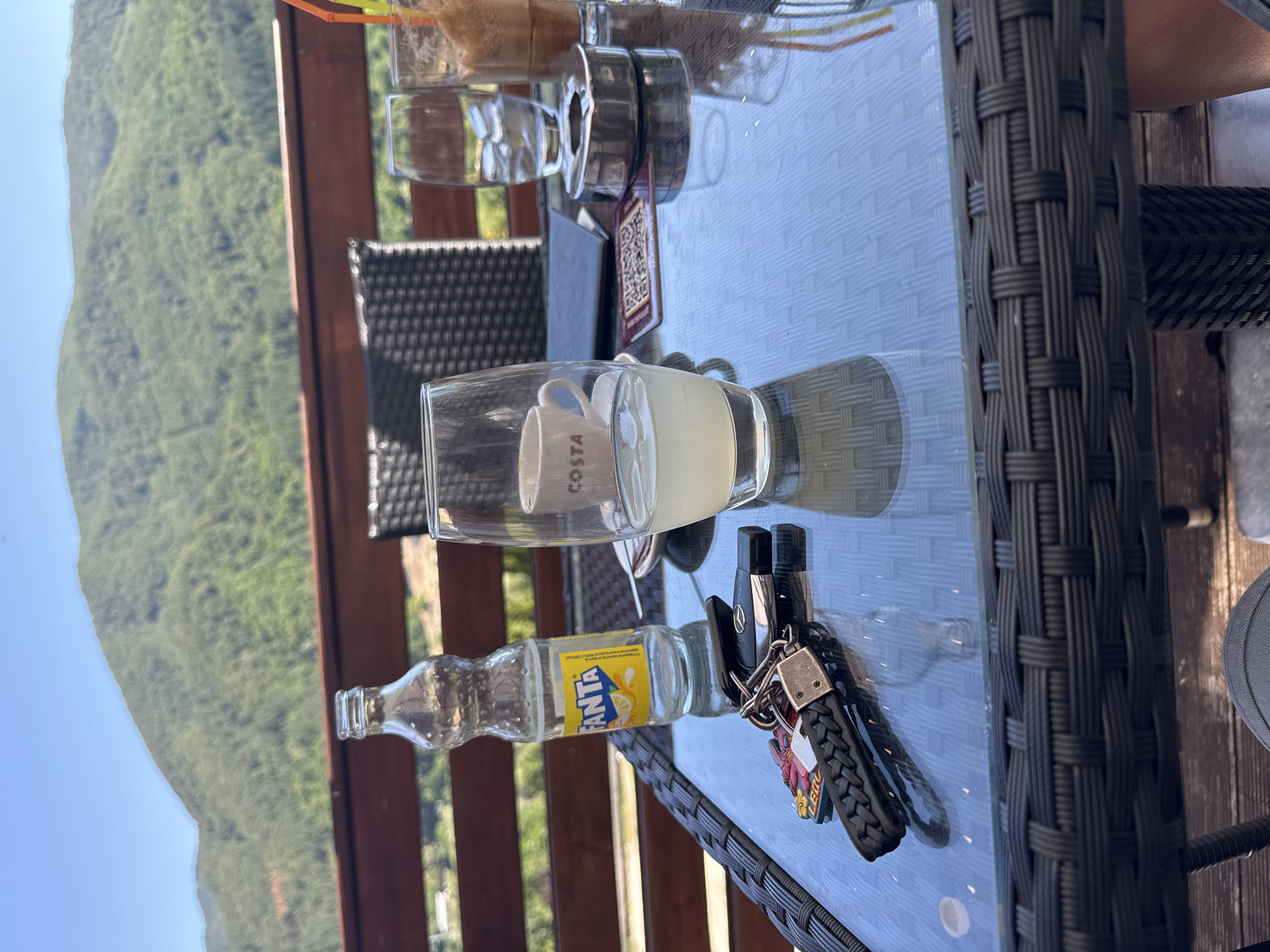 In Yurukovo, Bulgaria, a glass-topped table sits on a patio overlooking a lush, green mountainside under a clear blue sky. On the table, a half-empty bottle of Fanta, a glass of light-colored liquid with ice, a small Costa coffee cup, a metal ashtray, and a set of keys with a braided leather fob and a lobster charm are arranged. A second glass containing ice and a beverage with a straw is also visible. The table is surrounded by a woven outdoor chair and the scene appears to be set for relaxation and refreshment.