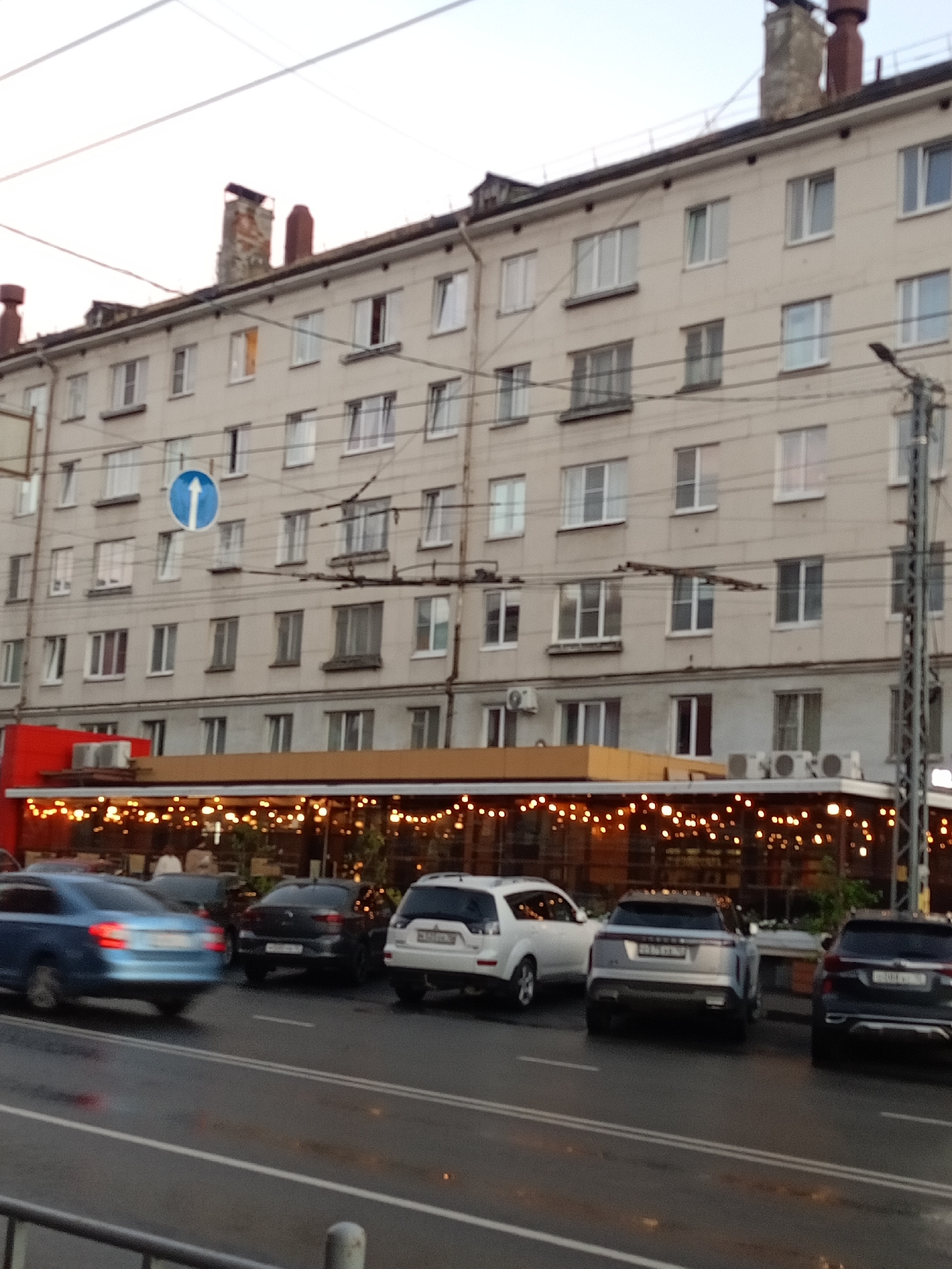 The image shows a street scene in front of a multi-story building. The building has a light-colored facade with numerous windows. A red-colored structure is attached to the lower part of the building on the left, and a covered outdoor seating area with string lights is visible in front of it. Several cars are parked and driving on the wet street. On the left side of the image, a blue car is moving away from the camera. A dark-colored SUV and a silver SUV are parked closer to the building. A dark-colored SUV is parked on the right side of the image. Overhead, power lines and a traffic sign with an arrow pointing upwards are visible. The sky is overcast, and the overall lighting suggests it is either early morning or late evening. The scene appears to be in a residential or commercial area.