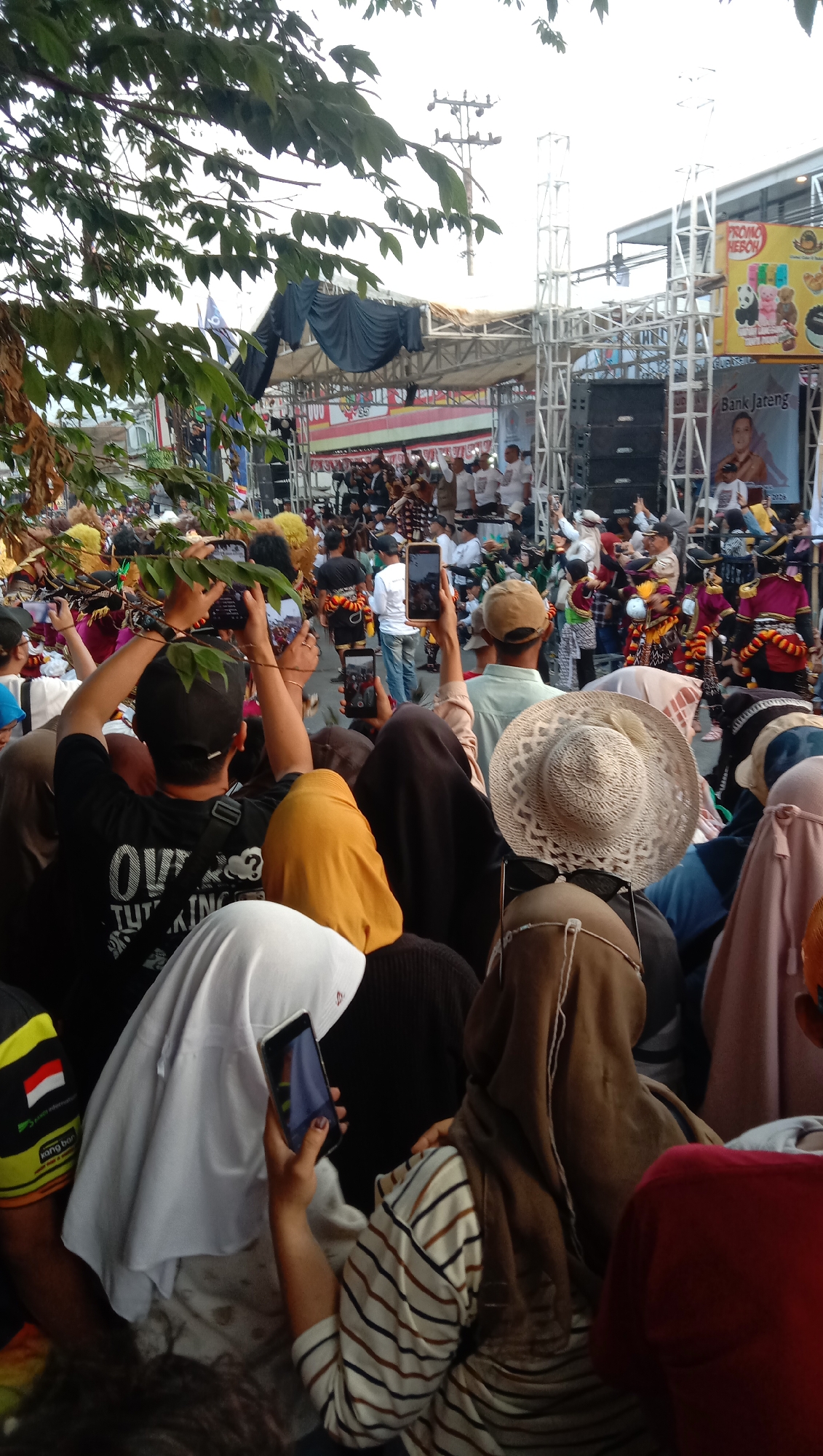 An outdoor daytime event is underway in Jatisawit, Indonesia, featuring a stage with musicians and performers. A large crowd of people are gathered, many of whom are holding up their mobile phones to record the event. A black draped structure hangs from a metal scaffolding above the stage. Banners promoting "PROMO HEBOH" and "Bank Jateng" are visible on the right side of the stage. The atmosphere appears to be festive, with a significant number of attendees wearing traditional attire and head coverings. Trees with green leaves frame the top of the image.
