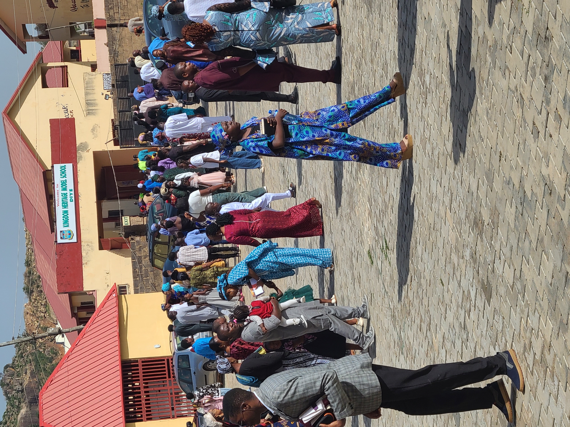 A daytime outdoor scene shows a large group of people standing on a paved area near a building, presumably in Unknown, Unknown. The building, possibly a school, has a sign that reads "KINGDOM HERITAGE MODEL SCHOOL DIYE." Most people in the group are dressed in casual or semi-formal attire. Some people are holding books or water bottles. Several cars are parked alongside the crowd. In the background, a small hill can be seen under a clear sky.