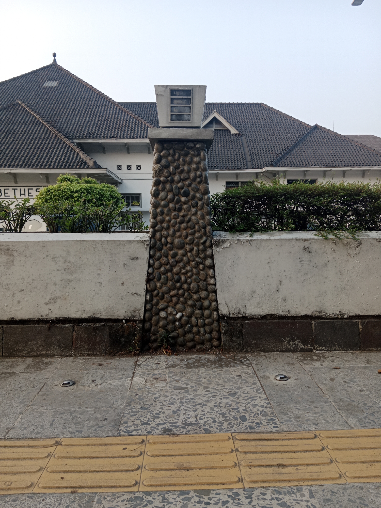 The image shows a low-angle, outdoor, daytime view of a building facade and a stone pillar in front of it. The building, likely a historic structure, has a dark, tiled, gabled roof and white walls with dark-trimmed windows and decorative "X" shaped motifs. Some text, partially visible, reads "BETHES". In front of the building is a low, rough concrete wall. A tall, cylindrical pillar made of small, rounded stones is positioned centrally in the foreground, against the concrete wall. The pillar has a light-colored, rectangular structure at its top, containing a barred window. The foreground consists of a sidewalk with textured yellow tactile paving tiles for the visually impaired, and grey concrete tiles with a speckled pattern. Several small, circular metal fixtures are embedded in the grey tiles. Lush green bushes are visible behind the concrete wall, to the left and right of the stone pillar. The sky is a pale, overcast blue. The scene appears to be located in a public area, possibly near a street or walkway. This image is of the Bethelehem Church in Yogyakarta, Indonesia.