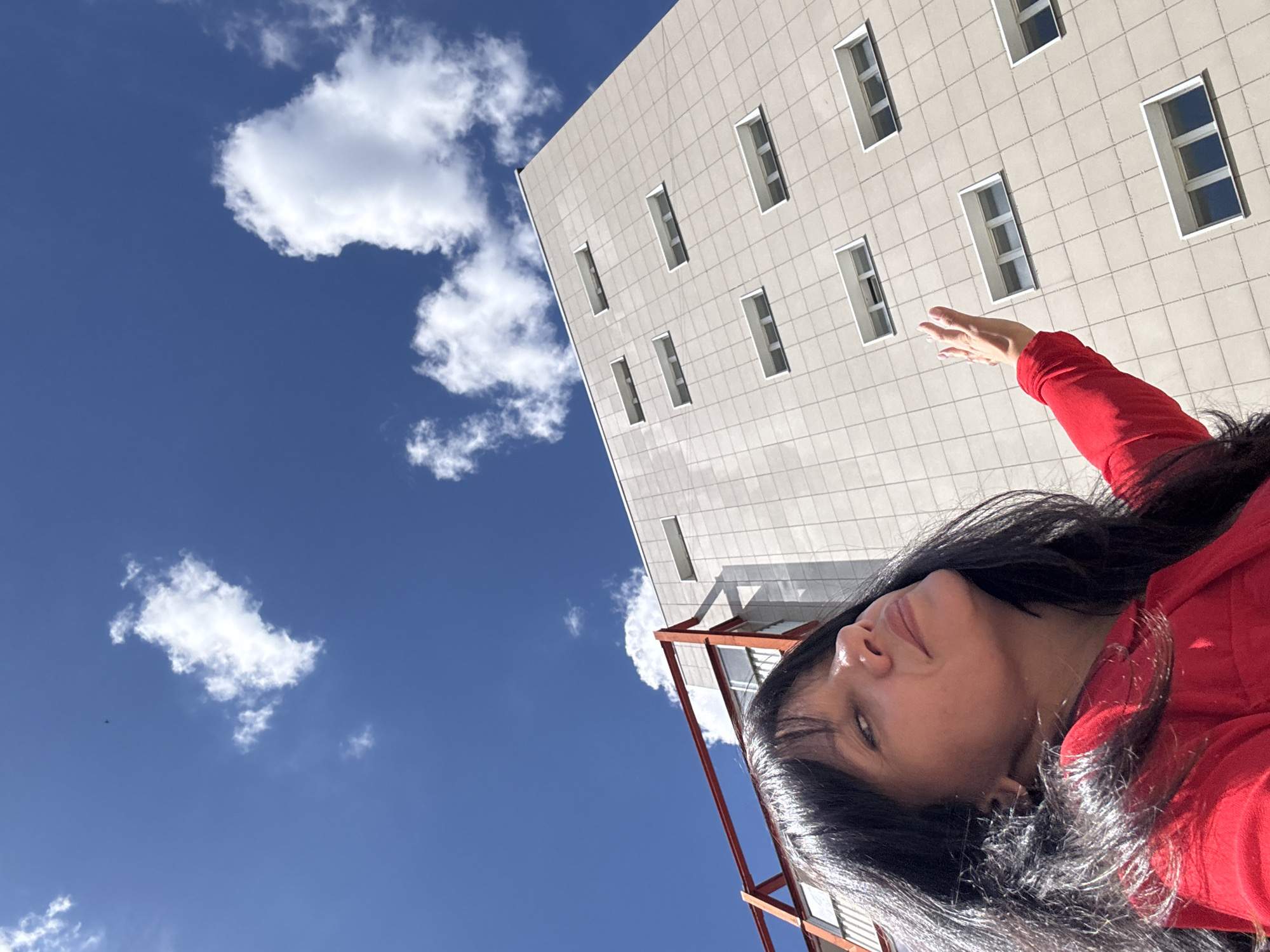 A woman in a red top looks up at a light-colored building with rows of rectangular windows against a blue sky with scattered white clouds. She is positioned in the lower left of the frame, with her face partially obscured by her dark hair. Her right arm is extended upward, palm facing outward, towards the building. The building is tall and appears to be a modern structure with a grid-like facade. A section of a red structure is visible on the left side of the building, possibly a balcony or architectural element. The scene is set outdoors during the daytime. There is no information about the location in Chaikovsky, Russia.