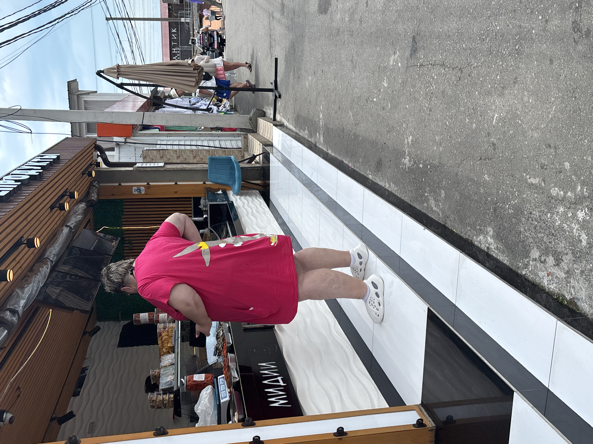 A person stands outside a food stall in Sochi, Russia, near the Black Sea. The stall, which sells seafood, is marked with the Russian word "МИДИИ" (mussels) and has an awning with lighting. The person, wearing a bright pink t-shirt with a cartoon character on it, and white clogs, is looking at the food display. Other shoppers and vendors are visible in the background, along with the typical street scene of a resort town, including a motorcycle and an umbrella. The weather appears to be partly cloudy.