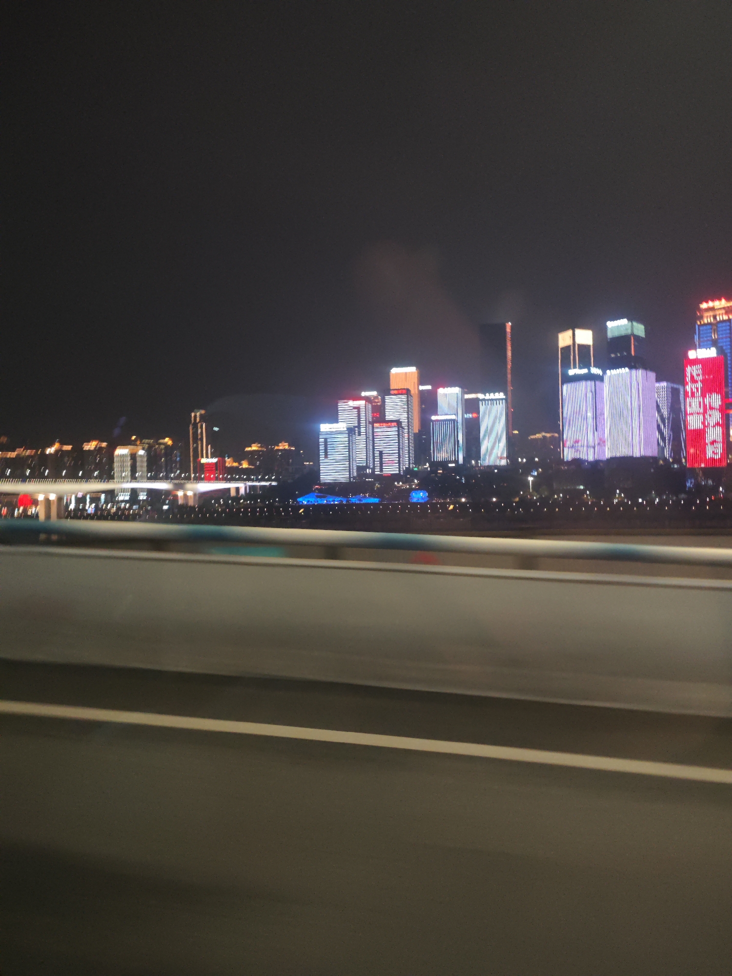 The image shows a nighttime cityscape, likely taken from a moving vehicle. The city is identified as Yuzhong District, China, based on the visual characteristics of the skyline. The skyline is dominated by illuminated skyscrapers with various lighting schemes, including white, red, and blue. A bridge or elevated roadway is visible in the foreground, separating the viewer from the city. A blur effect indicates the photo was taken while in motion. The sky is dark, with some cloud cover visible above the buildings.