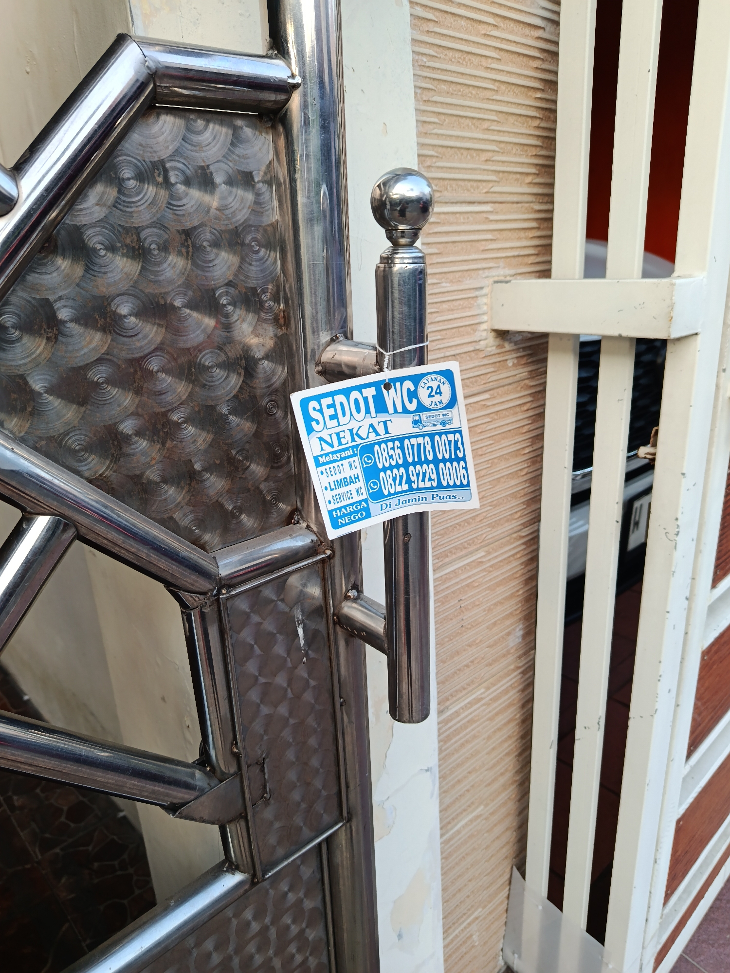 The image is a close-up shot of a metal gate with a blue and white sticker attached to one of its vertical posts. The sticker contains text and icons advertising a toilet service. The text on the sticker reads: "SEDOT WC NEKAT", "Melayani : •SEDOT WC •LIMBAH •SERVICE WC", "HARGA NEGO", and "Di Jamin Puas..". There are also phone numbers listed: "0856 0778 0073" and "0822 9229 0006". The background shows a textured wall and a portion of another fence or gate. The image appears to be taken outdoors in Keboananom, Indonesia, a location known for its historical significance.