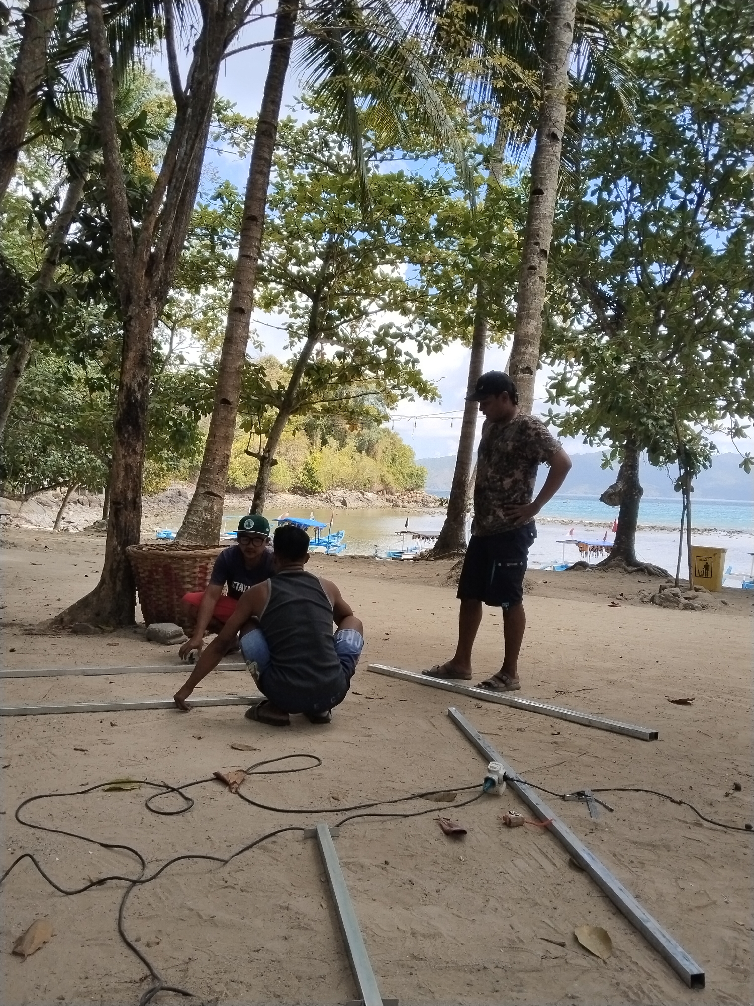 Three men are on a sandy beach, likely in Prigi, Indonesia, working with metal poles. One man, wearing a camouflage shirt and black shorts, stands with his hands on his hips, observing the others. Another man, in a dark vest and blue shorts, kneels, appearing to work on the metal poles. A third man, wearing a dark t-shirt and red shorts, is also kneeling and seems to be assisting with the metalwork. The ground is sandy with scattered dry leaves. Several metal poles are laid out, some connected, with electrical cords and what appears to be a welding apparatus visible on the ground. In the background, there are several palm trees and other lush vegetation. Beyond the trees, a body of water with several small boats is visible, suggesting a coastal location. The sky is partly cloudy.
