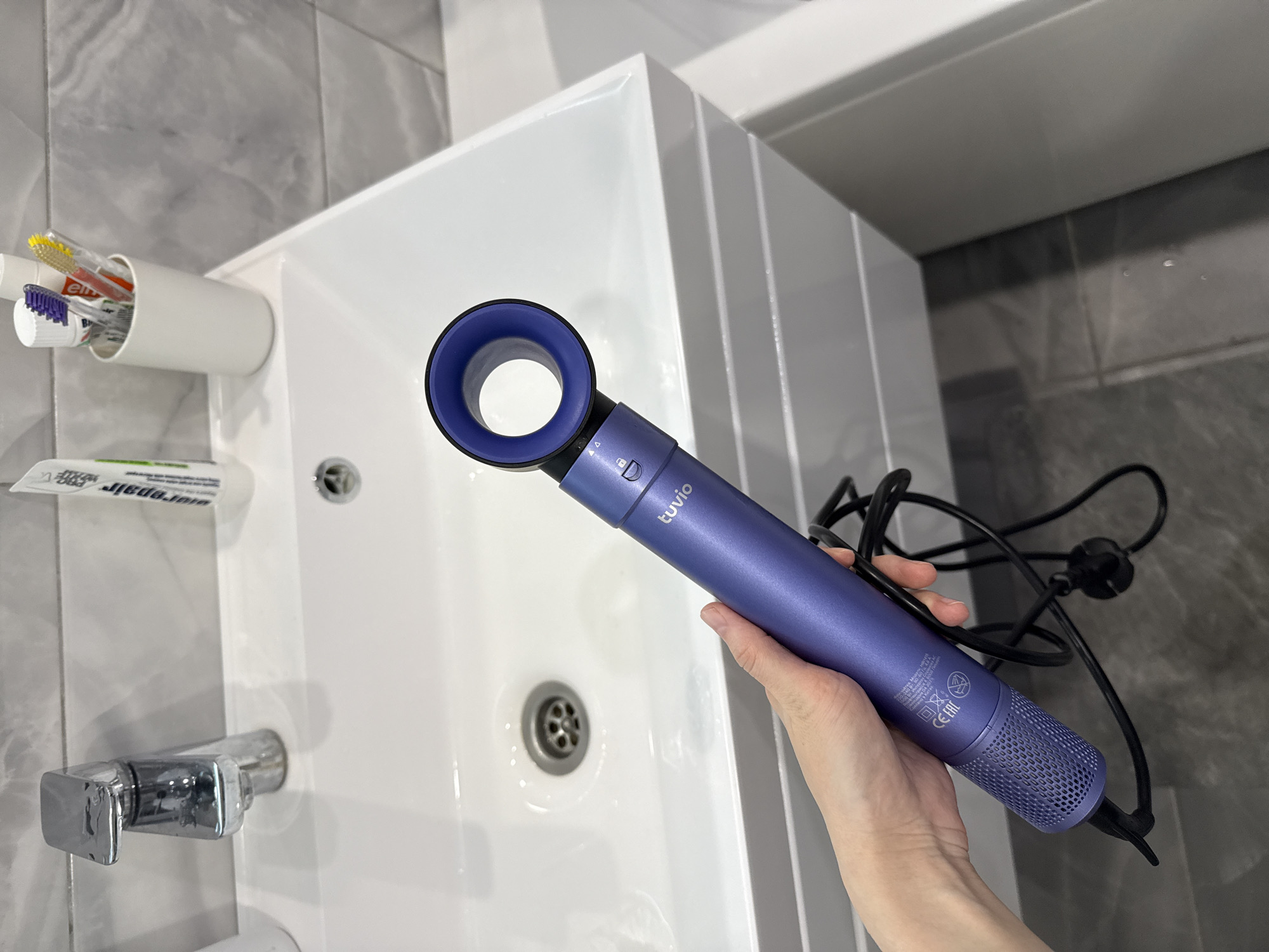 A person is holding a purple Tuvio hairdryer over a white bathroom sink. The sink has a silver faucet on the left side and a drain in the center. To the left of the sink is a white container holding toothbrushes and a tube of toothpaste. The background shows gray and white tiles on the wall and a white cabinet above the sink.