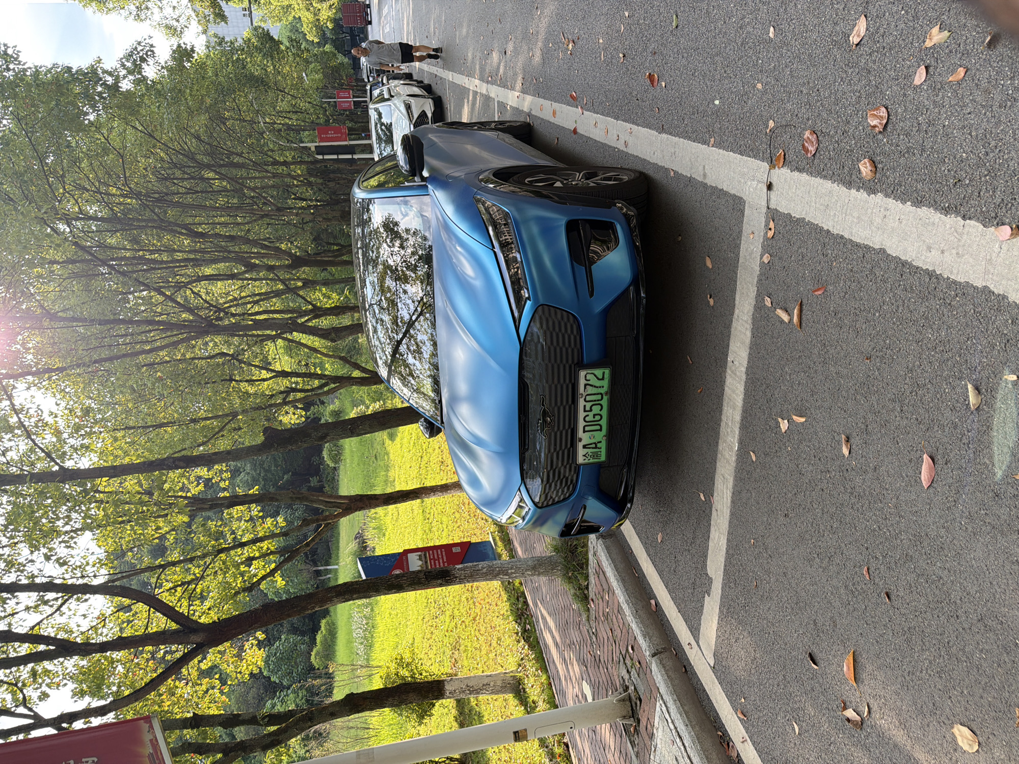 The image shows a blue Ford Mustang Mach-E parked on a street in Jiangjin, China. The car is in the foreground, facing slightly to the right. It has a green license plate that reads "A·DG5072". Behind the car, a line of other vehicles is parked along the street, and a man in a grey t-shirt and black shorts is walking away from the camera. The street is lined with trees, and there is a grassy area with sunlight filtering through the leaves. Fallen leaves are scattered on the asphalt. The overall environment suggests a bright, sunny day in a park-like setting within the city.