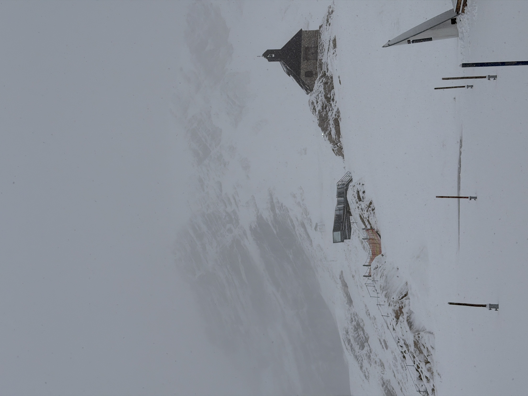 The image depicts a desolate, snowy mountain landscape during a snowstorm, likely at the Zugspitze, Germany's highest peak, near Garmisch-Partenkirchen. In the foreground, wooden posts mark a path or boundary in the snow. A modern glass structure, possibly an observation deck or part of a ski resort, is visible on the left side of the image, connected to a pathway with railings and orange fencing. Further up the slope, on a rocky outcrop, stands a small, stone chapel with a dark, steeply pitched roof and a bell tower. The surrounding mountains are shrouded in thick fog and snow, making visibility poor. The overall atmosphere is stark and wintry, with visible snowflakes in the air. There are no people visible in the image.