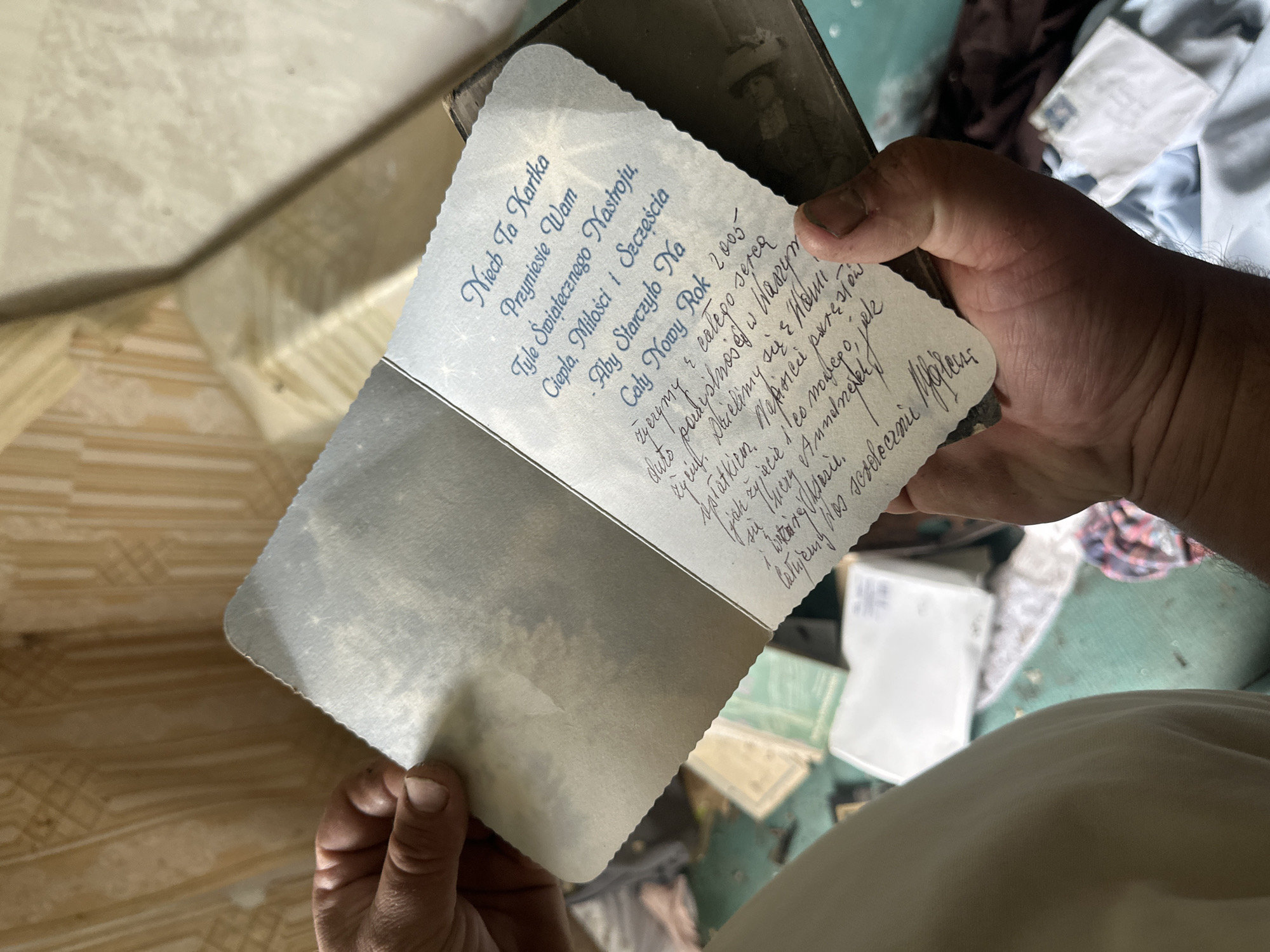 An individual's hands hold open a handwritten card. The card is light blue with a scalloped edge and features printed text in blue script at the top, followed by handwritten cursive below. The printed text reads, "Niech Ta Kartka Przyniesie Wam Tyle Świątecznego Nastroju, Ciepła, Miłości i Szczęścia". Below this, the handwritten text begins, "Aby Starzyło Na Cały Nowy Rok 2005". The remainder of the handwritten text is difficult to decipher fully due to the angle and resolution, but appears to be a personal message. The background suggests an interior space with weathered walls and scattered items on the floor, hinting at neglect or abandonment. The scene is likely in a residential setting, possibly an abandoned home, located in the Відзаўскі сельскі Савет, Belarus, as indicated by the prompt. The focus is on the card and the act of holding it.