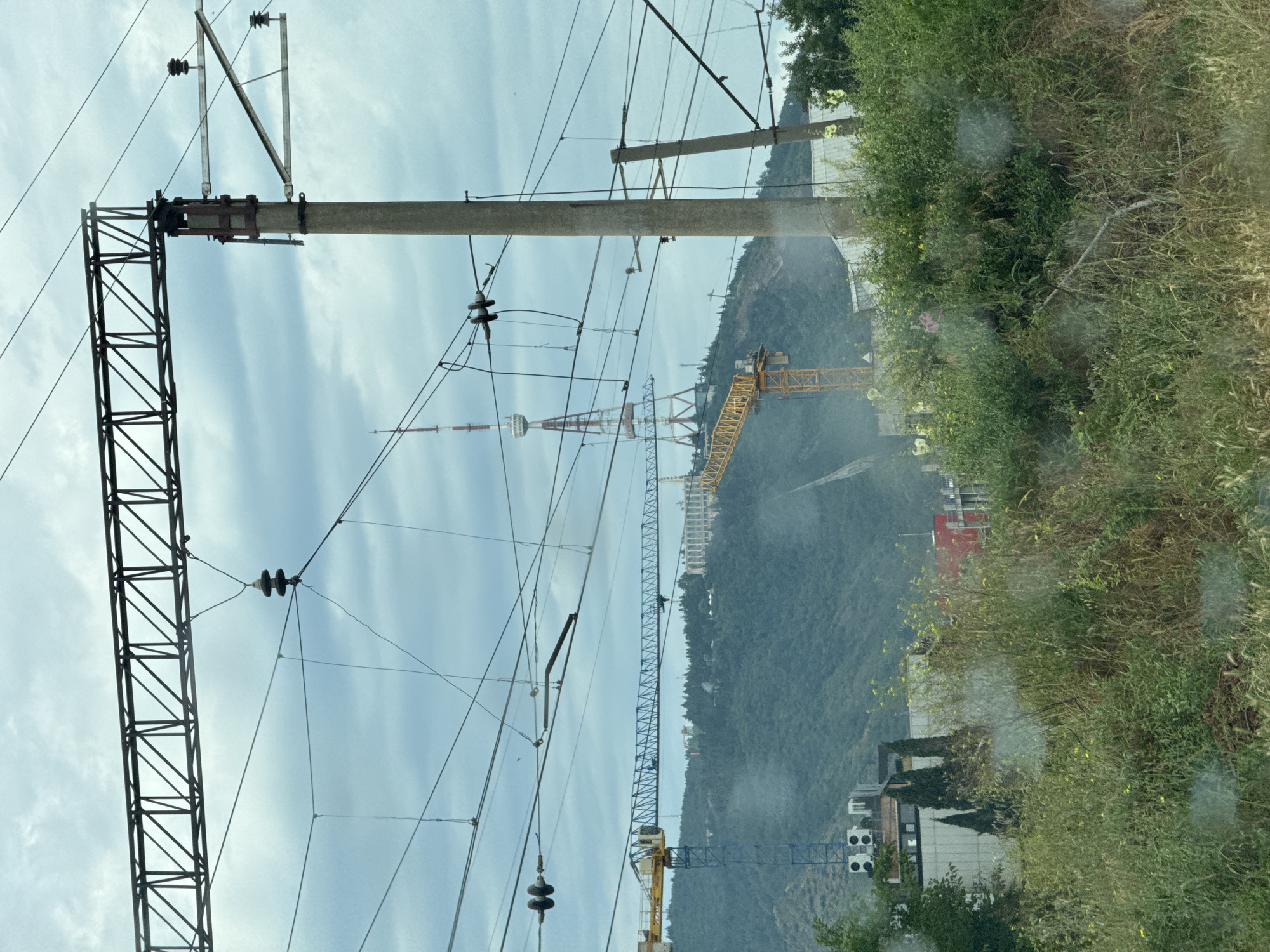 This image depicts a view from a moving vehicle in Tbilisi, Georgia, characterized by overhead power lines and electrical infrastructure in the foreground, suggesting a transit corridor such as a railway line. In the mid-ground, the hillside is dotted with trees and features construction activity, including two large yellow cranes and a white building with blue accents. Dominating the background is the Tbilisi TV Tower, a prominent landmark situated on a hill, and a large white building complex on the same hillside. The sky is overcast with scattered clouds. The vegetation in the immediate foreground is dense and overgrown.
