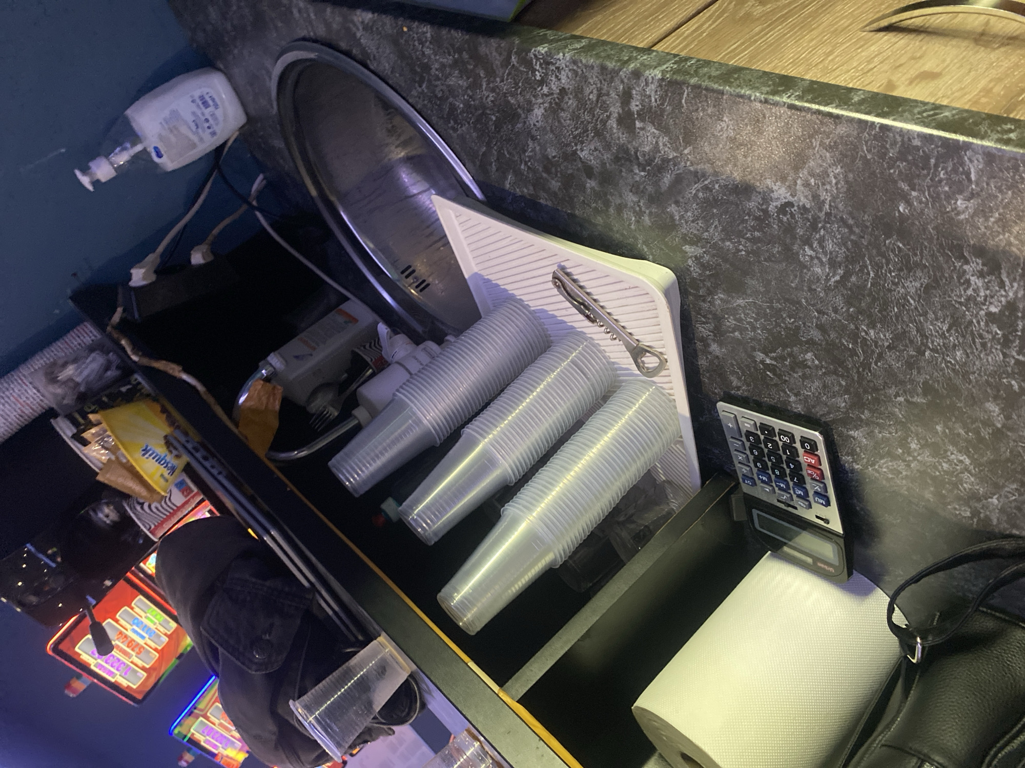 The image depicts a cluttered countertop area, likely in a commercial or service establishment, with a backdrop of slot machines, suggesting a casino or entertainment venue. The countertop itself is a dark, speckled material. Prominently featured are stacks of clear plastic cups arranged in a holder. A bottle opener is placed on the cup holder. To the left, a calculator and a roll of white paper are visible, along with a black handbag. Further back, a bottle of liquid soap is present, along with some electrical cords and outlets. The overall impression is one of a utilitarian space with items for dispensing drinks and possibly for cleaning. The scene appears to be indoors, with artificial lighting. No people are visible in the image. The image's context within Beloslav, Bulgaria, is not discernible from the visual information alone.