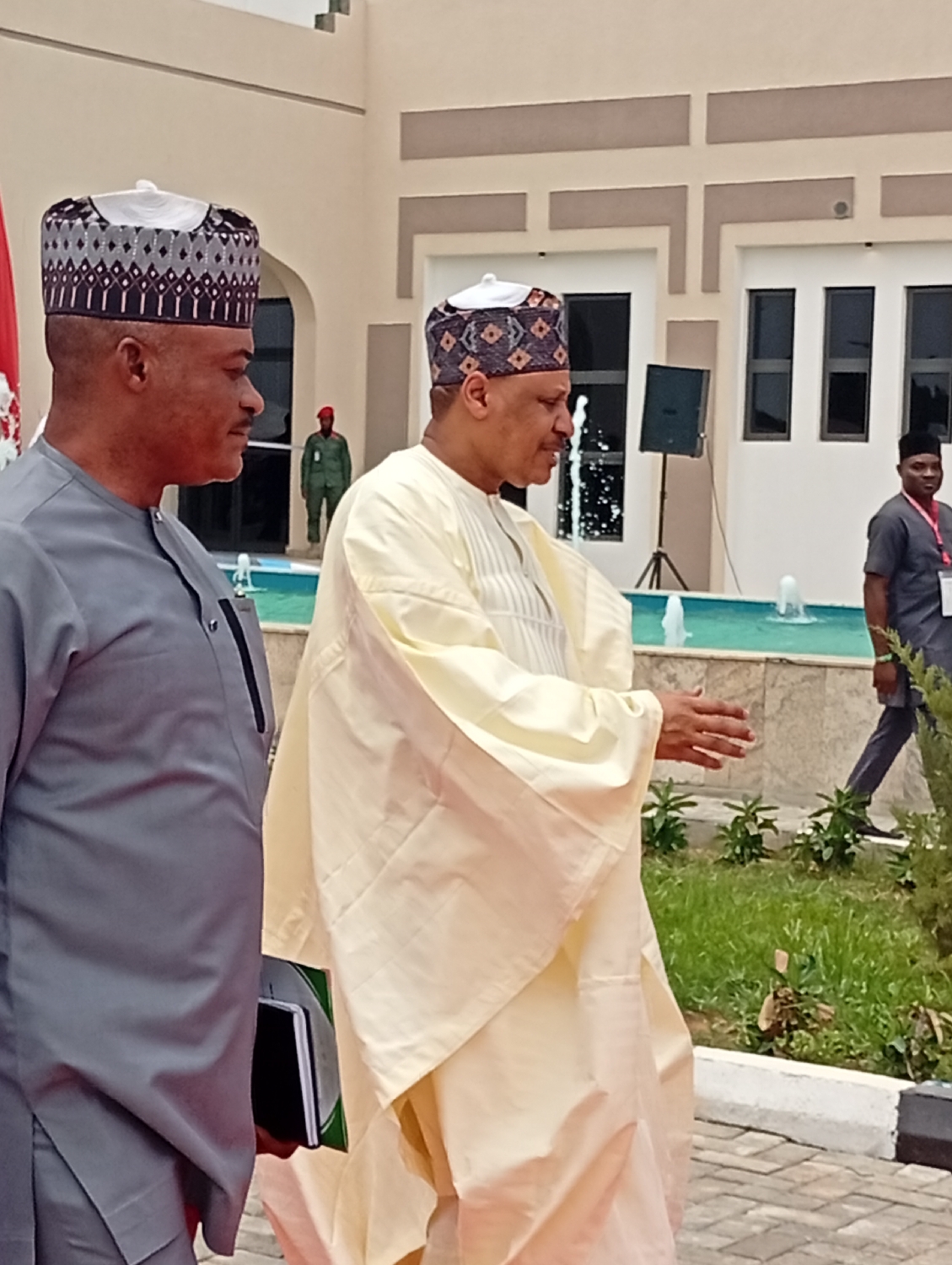 The image captures a bright, daytime scene at what appears to be a formal outdoor event in Unguwan Sanusi, Nigeria. Two prominent men, likely dignitaries, walk from left to right in the foreground. The man on the left wears a grey traditional tunic (agbada or kaftan) and a patterned grey and white kufi hat. He holds a dark notebook in his left hand. The man on the right wears a cream or light yellow traditional tunic and a similarly patterned dark and white kufi hat, actively gesturing with his hands as if speaking or explaining.
They are positioned on a paved surface, possibly a red carpet, with green grass and foliage to their right. Behind them, a decorative fountain with active water jets adds to the scene. In the mid-ground, a modern building with light-colored walls and dark rectangular windows forms the backdrop. A black speaker on a tripod stand is visible near the fountain.
Several other individuals are present in the background. To the far left, partially obscured, is a person in a red hat and a green military-style uniform. To the far right, a man in a dark shirt and trousers, wearing a badge, walks away from the camera. The overall setting, including the traditional attire, the formal architecture, the red carpet, and the presence of what appears to be security personnel, suggests an official meeting, reception, or public event. The clear skies indicate good weather. No discernible text is visible in the image.