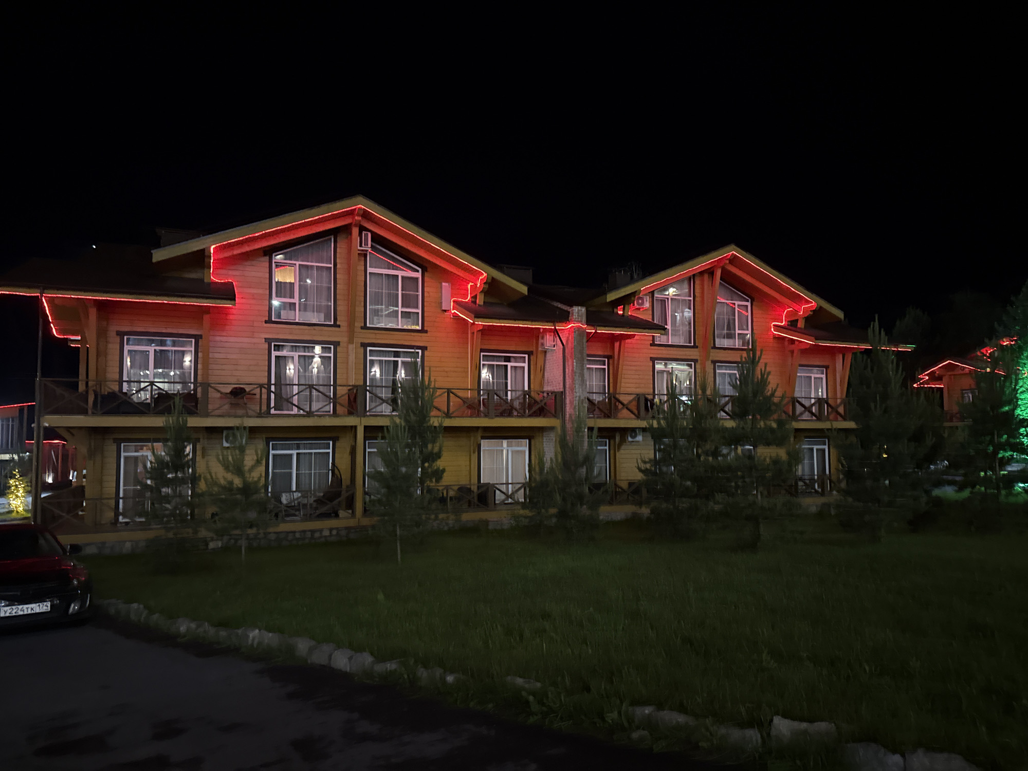 This is a night-time, horizontal, eye-level shot of a modern, multi-story wooden building with red lights outlining its roof and balconies. The building appears to be a resort or hotel, with multiple windows and balconies visible. The structure is made of horizontal wooden planks and features a dark-colored roof. Red LED lights are prominently displayed along the eaves and edges of the balconies, creating a festive or decorative effect. In front of the building, there is a well-maintained lawn with several young evergreen trees. To the left of the building, a dark-colored car is parked on an asphalt surface. The sky is dark, indicating it is nighttime, and a few other illuminated structures are visible in the background to the right, one with green lights. The overall scene suggests a pleasant, illuminated outdoor setting at night, possibly a tourist destination. The image is located in the city of Syrostan, Russia.