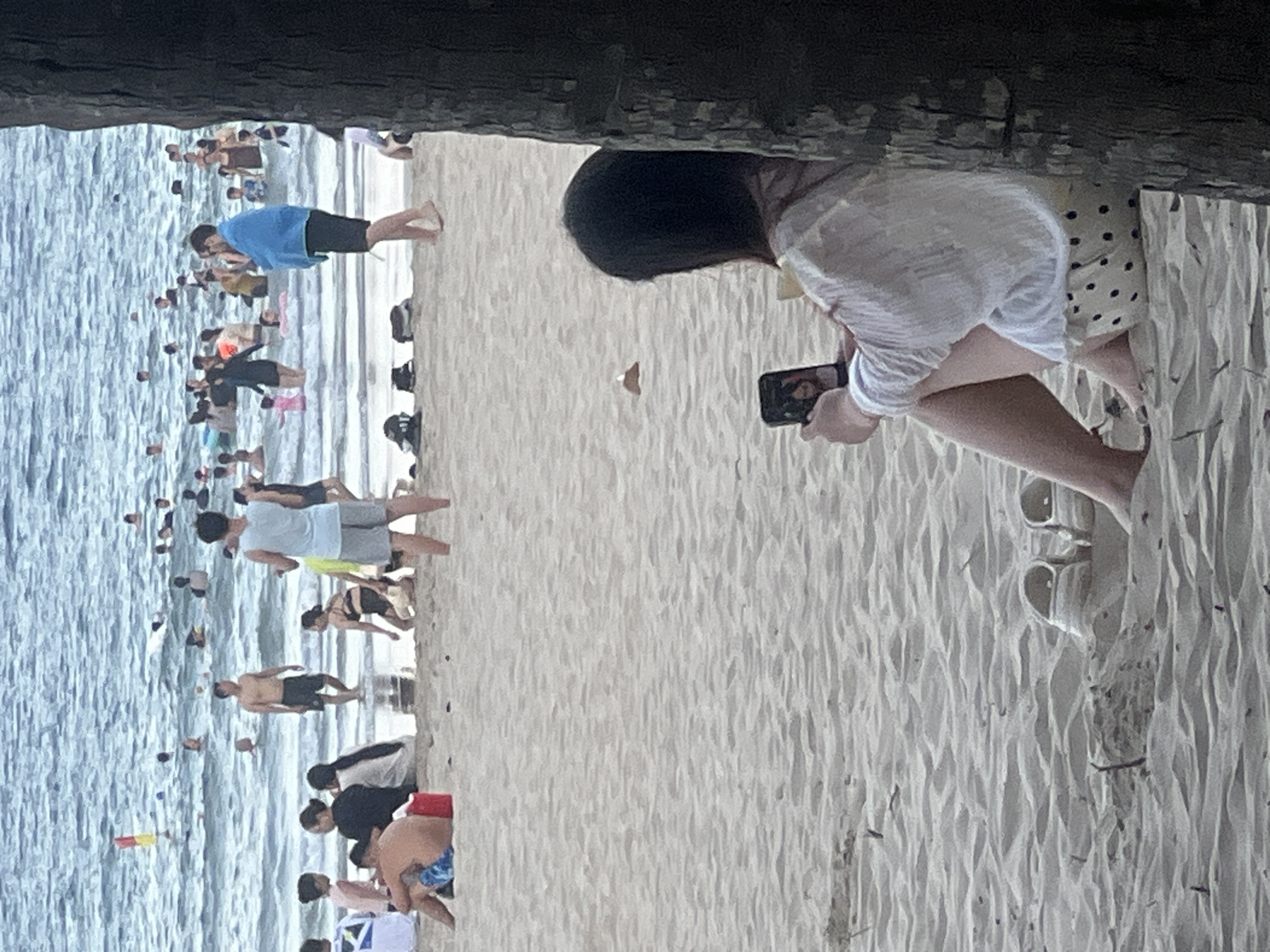 The image depicts a crowded beach scene in Da Nang, Vietnam. In the foreground, a woman with dark hair sits on the sand, looking at her phone. She is wearing a white, loosely fitting shirt and a patterned skirt. Nearby, a pair of white sandals are partially buried in the sand. The middle ground shows a variety of people on the beach, some sitting and some standing. Further back, the ocean is filled with numerous swimmers and people wading in the water. A red and yellow buoy is visible in the water on the left side of the image. The overall impression is of a popular beach on a busy day.