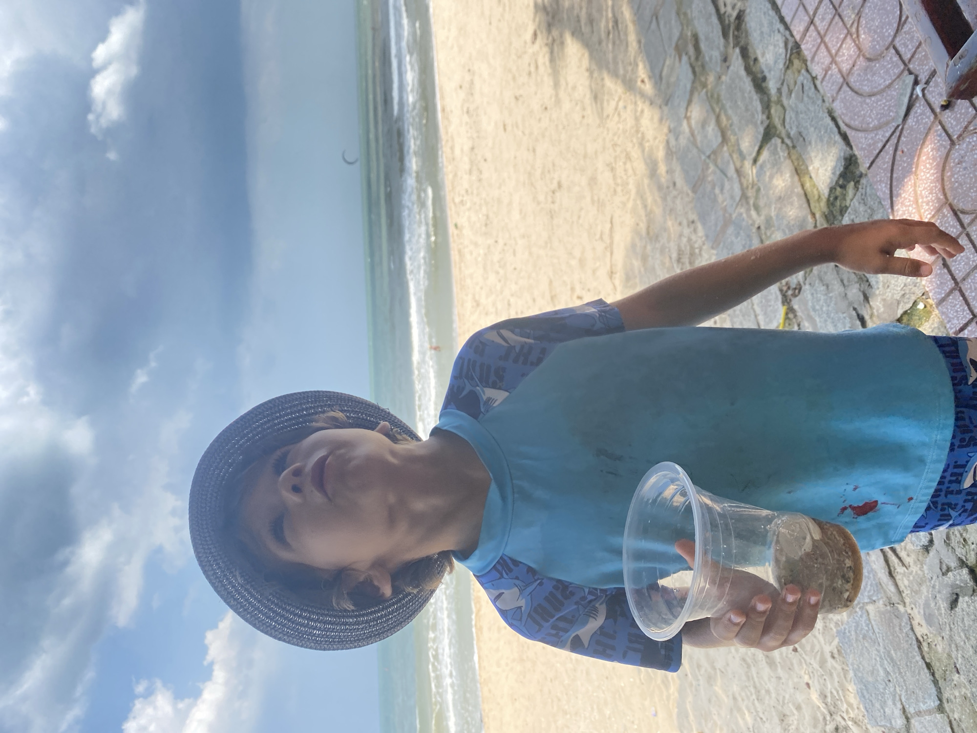 A young boy wearing a blue rash guard and a dark hat stands on a beach in Mũi Né, Vietnam. He is looking upwards and holding a clear plastic cup containing a dark liquid and some solid debris. The beach sand is light brown, and the ocean with waves is visible in the background. The sky is cloudy, with some lighter patches. A kite surfer can be seen in the distance on the water. The boy is positioned on a paved area with stone tiles and a tiled border.