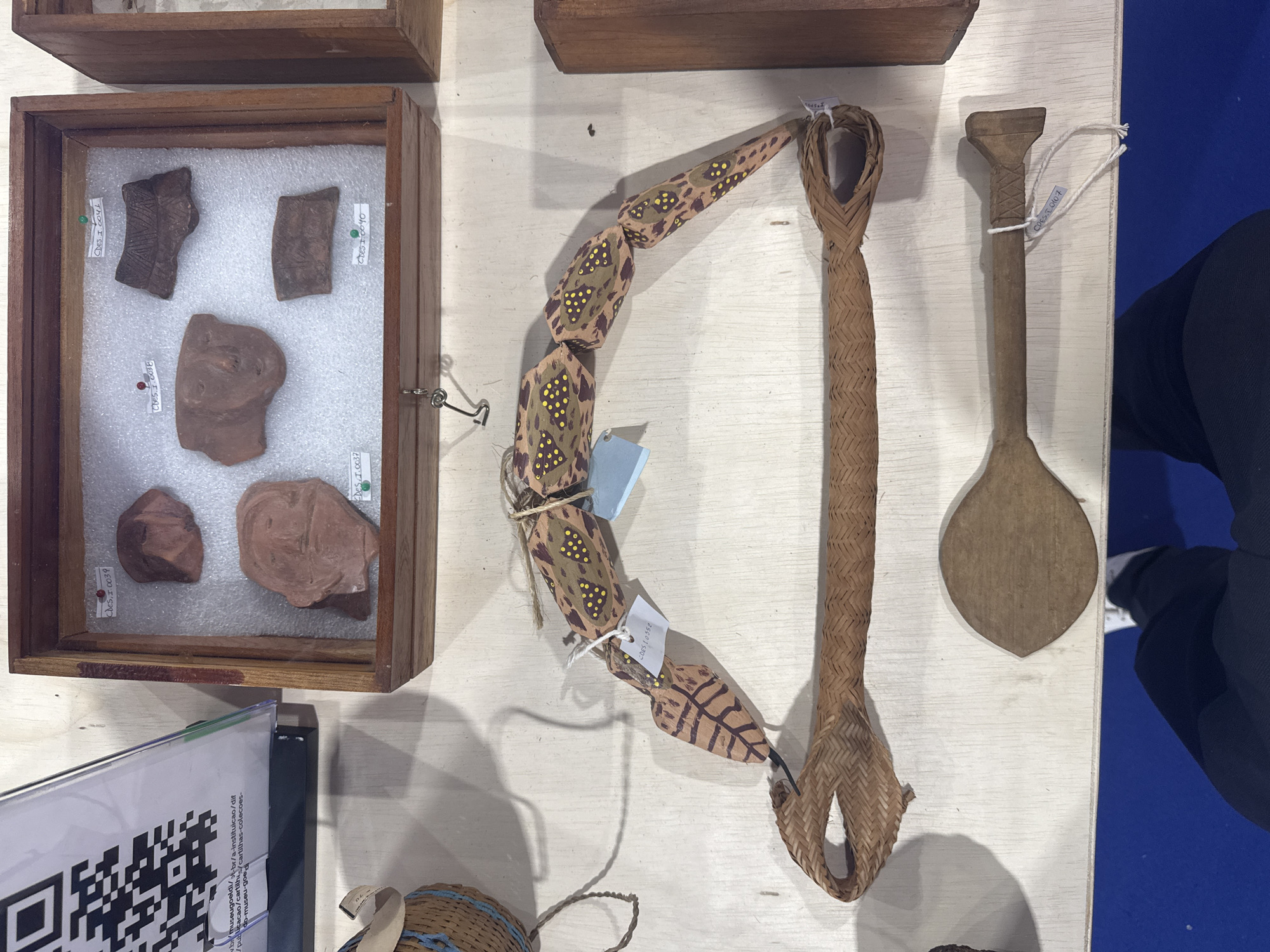 The image displays an exhibition of indigenous artifacts on a white wooden surface. In the upper section, a wooden box contains several clay fragments with labels such as "CDES.I.0039," "CDES.I.0041," "CDES.I.0038," "CDES.I.0037," and "CDES.I.0040." To the left of the box, a QR code is displayed on a slanted white board. Below the box, a segmented wooden object, painted with brown and yellow patterns, is suspended. Attached to this object is a white tag labeled "CDES.I.0352." Below the segmented wooden object is a woven brown artifact, possibly a handle or strap, and beneath that, a flat wooden object resembling a spoon or paddle. A small white tag with "CDES.I.0107" is attached to the wooden spoon-like object. In the lower portion of the image, a person's legs and feet are visible, wearing dark pants and white sneakers, standing on a blue surface. The overall scene suggests an indoor exhibition, likely in a museum or gallery in Niterói, Brazil, given the context of indigenous artifacts.