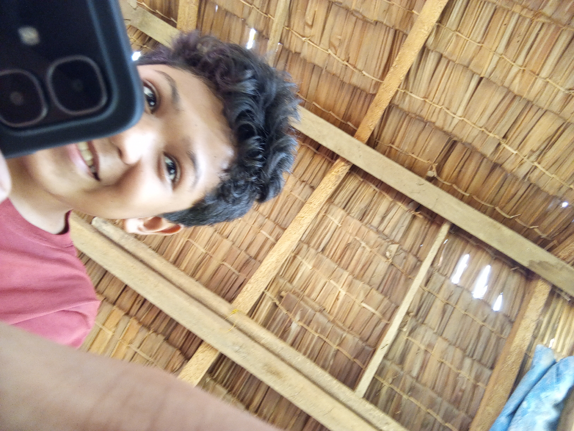 A person's face is partially visible, held up to a dark-colored smartphone, with the camera facing the user. The person is wearing a pink t-shirt and their dark, curly hair is visible. The background is a thatched roof made of dried leaves. The image is taken from a low angle looking up.
