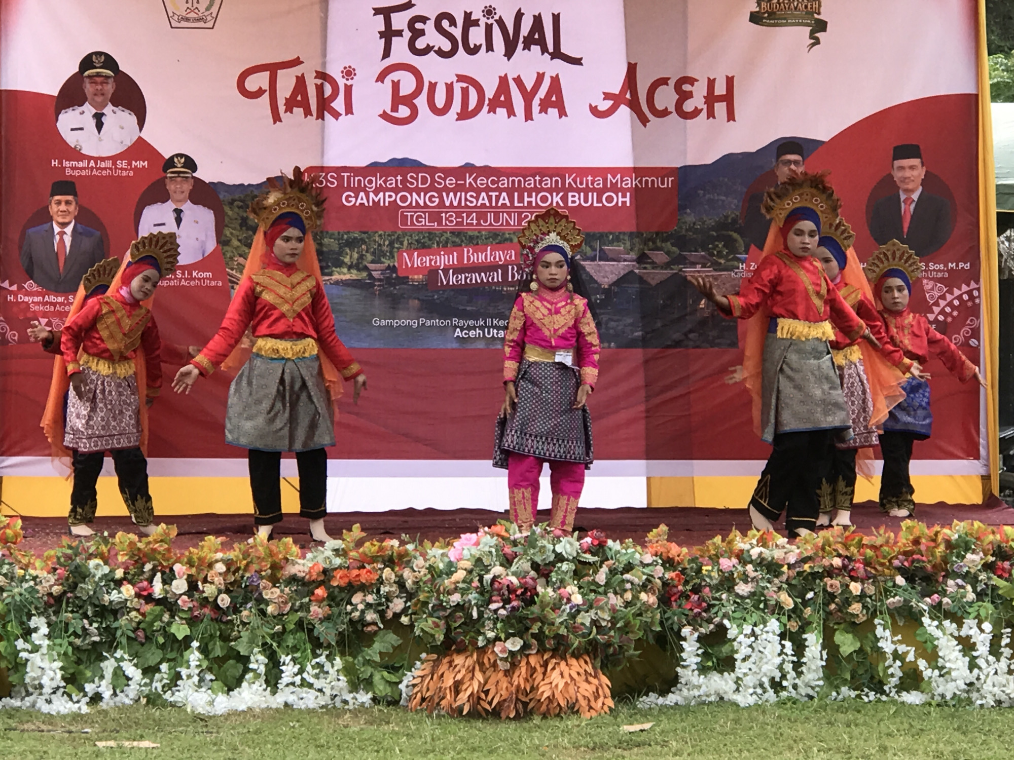 The image shows a stage decorated with flowers and foliage, set against a backdrop that reads "FESTIVAL TARI BUDAYA ACEH". Four young girls in traditional Acehnese attire are performing on stage. The girls are wearing vibrant red tops with gold accents, patterned skirts, and elaborate headpieces. Two of the girls are positioned in the foreground, with one on the left and another slightly to the right of center, while the other two are further back. The backdrop also features portraits of officials, indicating a cultural event or ceremony. The scene appears to be outdoors, with a grassy area in the foreground. The presence of the text "KOTA LANGKUTA" is not explicitly visible in the provided image or crops, so no assertion about the city of Langkuta can be made based solely on this visual information.