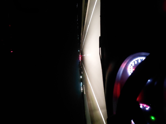 A nighttime perspective from inside a moving car. The illuminated dashboard displays a speedometer with red accents. A steering wheel is partially visible in the foreground. Outside, road markings and distant lights create a visual perspective. A highway lane is distinguished by white lines, leading to the glow of other vehicle lights. The scene is dark, highlighting the contrast between interior car lights and the external environment.