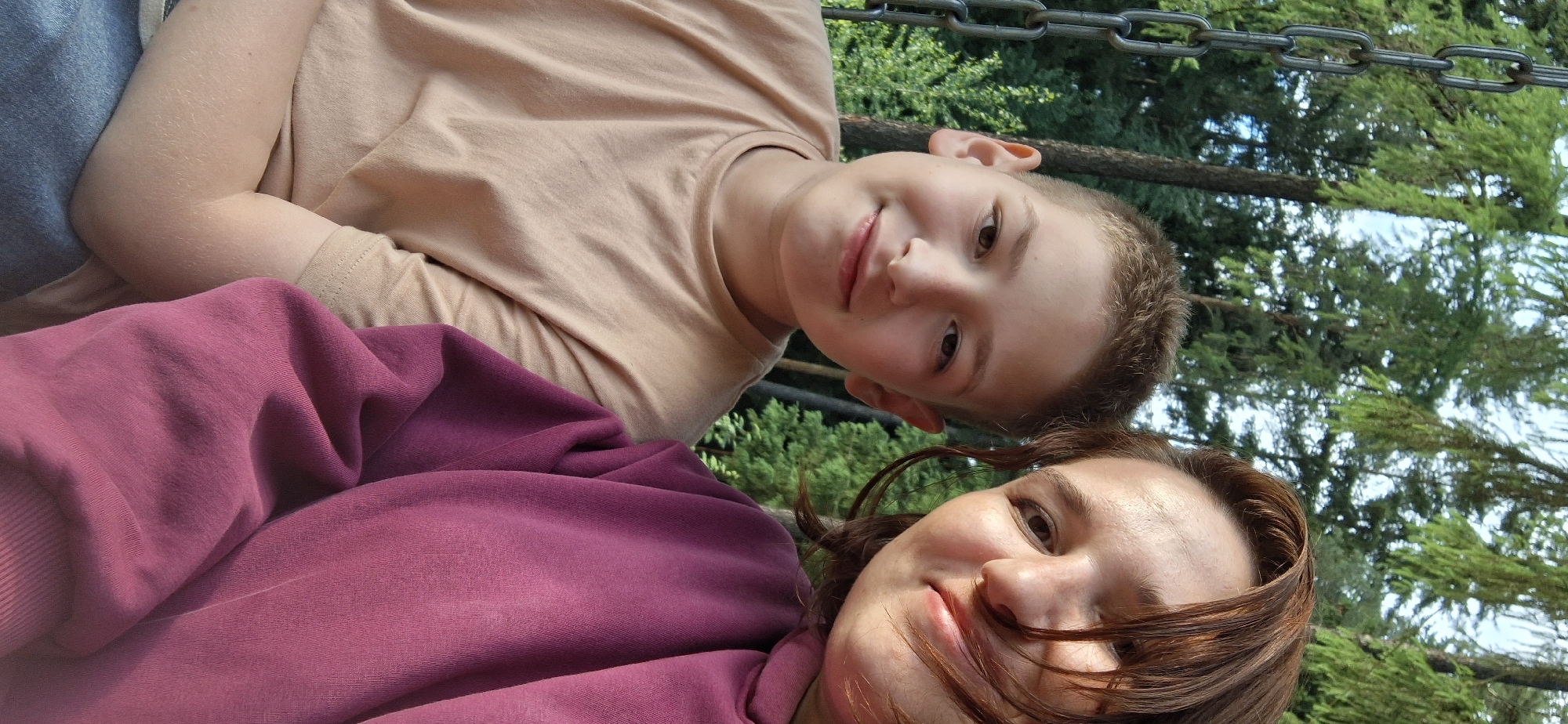 Two people, an adult and a child, are in the frame. The child, wearing a tan t-shirt, is positioned above the adult. The adult is wearing a maroon hoodie. They appear to be lying down outdoors, possibly in a park or natural area, indicated by the presence of green trees and a metal chain structure in the background. Based on the location information, this scene is likely located in Прикамский, Russia.