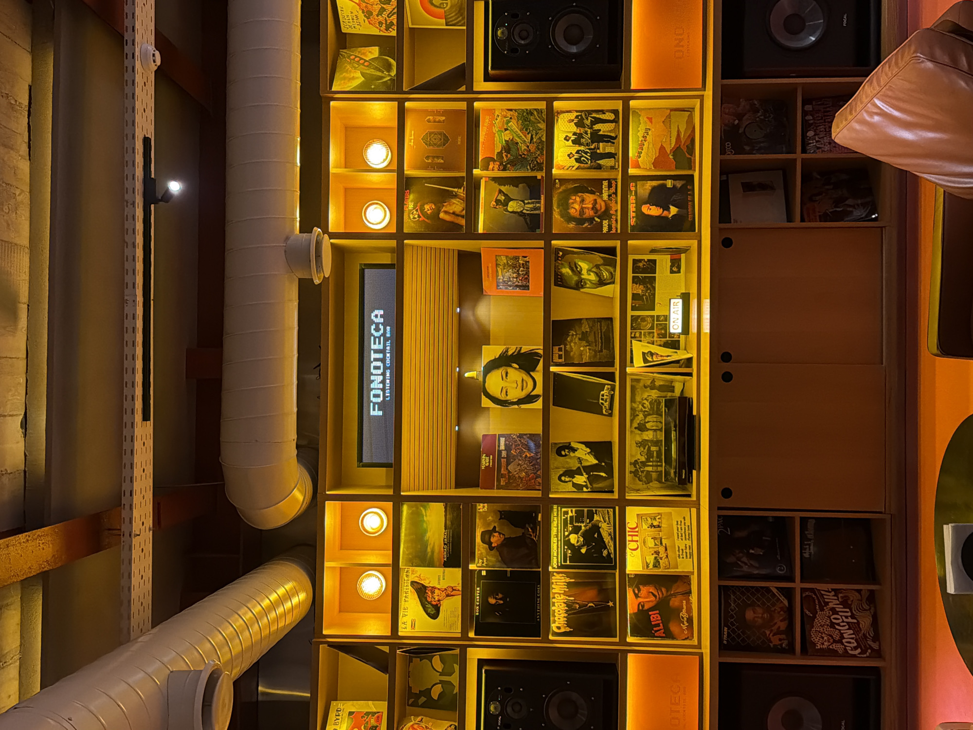 A view of the ceiling featuring a wall-mounted structure of shelves in Yerevan, Armenia. The shelves are filled with vinyl records, speakers, and small light fixtures. A digital display is positioned in the center, reading "FONOTECA LISTENING COCKTAIL BAR". Two parallel white pipes run vertically alongside the shelf structure. A metal rack is also visible next to the pipe.
