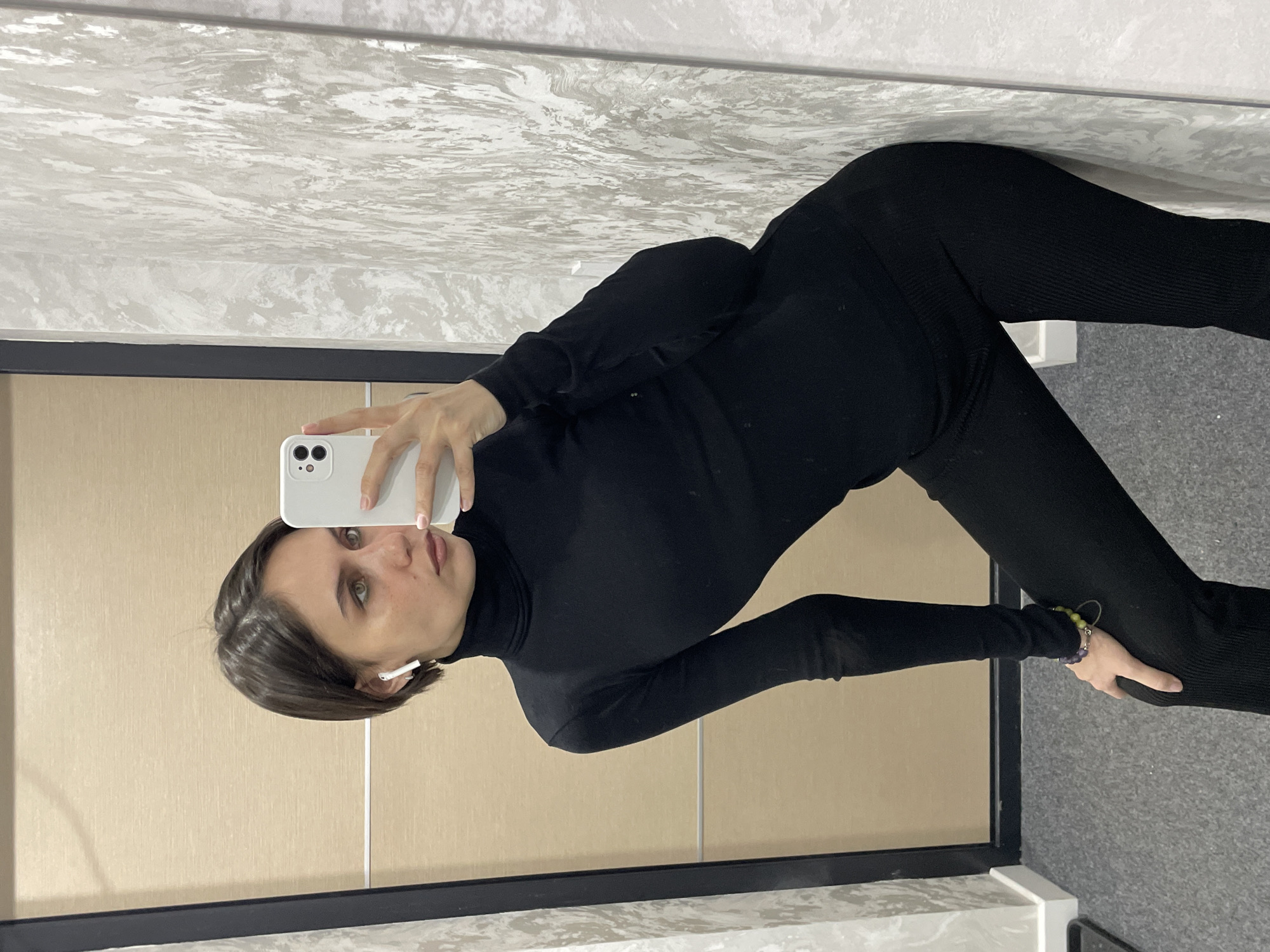 An indoor mirror selfie captures a female individual with short, dark brown hair and green eyes. She wears a black long-sleeved turtleneck and black ribbed pants. A white wireless earbud is present in her left ear, and a multi-colored beaded bracelet is visible on her left wrist. She holds a white smartphone with her right hand, extended to photograph her reflection. Her left hand rests on her bent left knee, and she is leaning slightly against a light gray and white textured wall. To the left, a light beige wood-paneled door with a black frame is visible. The floor is covered in gray carpet. The precise location within Izhevsk, Russia, cannot be determined from the provided visual information, other than being an interior space.