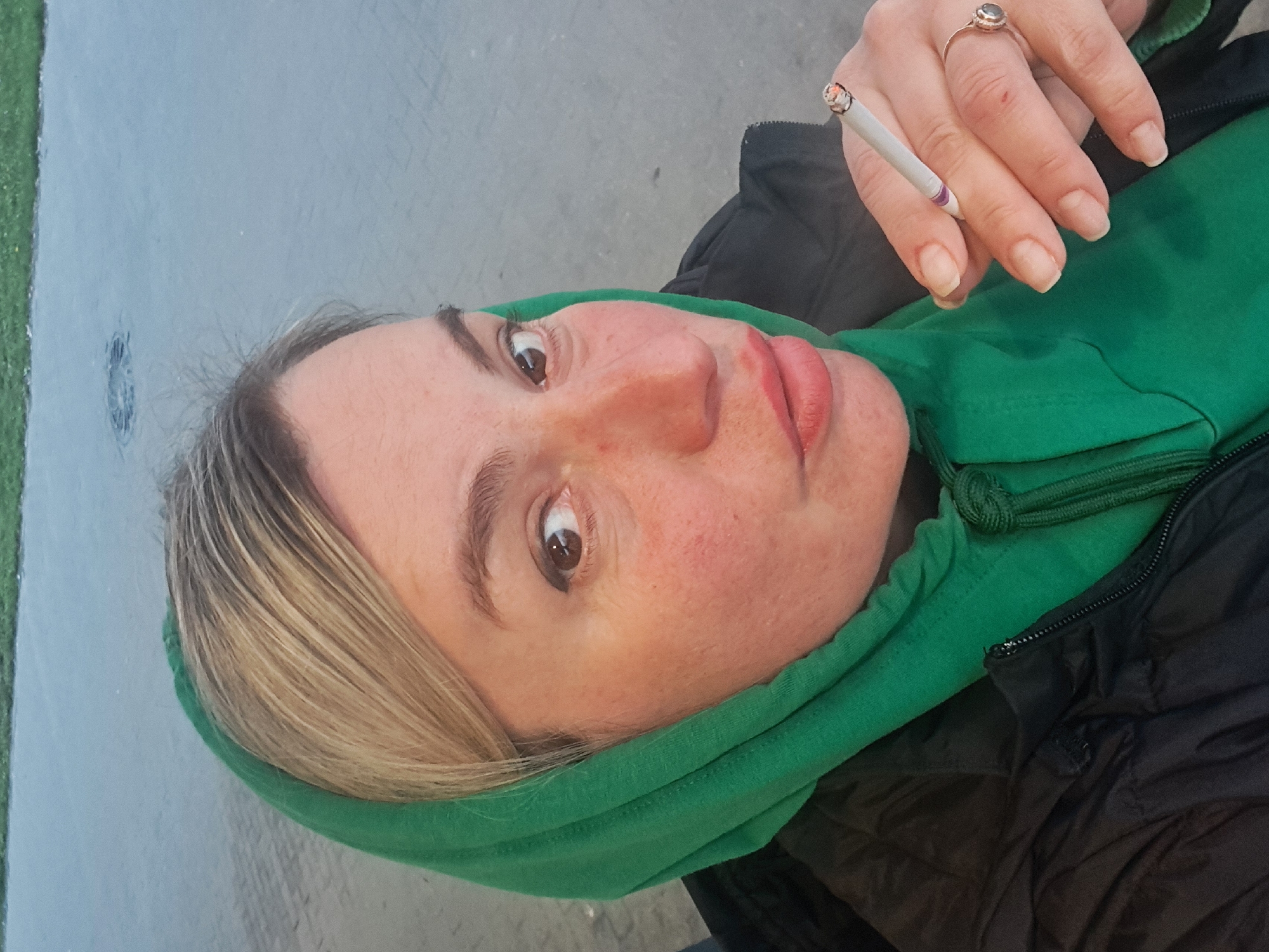 A woman wearing a green hooded top and a black jacket is holding a lit cigarette between her fingers. She is looking directly at the camera. She is wearing a ring on her finger. The location is not discernible as Elektrostal, Russia. The background appears to be a gray surface, possibly concrete or asphalt.