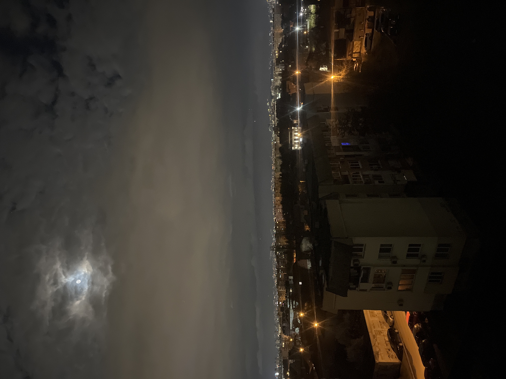 An eye-level, long outdoor exposure taken at night depicts a city at night in Sofia, Bulgaria. The sky is cloudy with a visible moon. Buildings and streetlights illuminate the lower right, and the city extends into the distance.