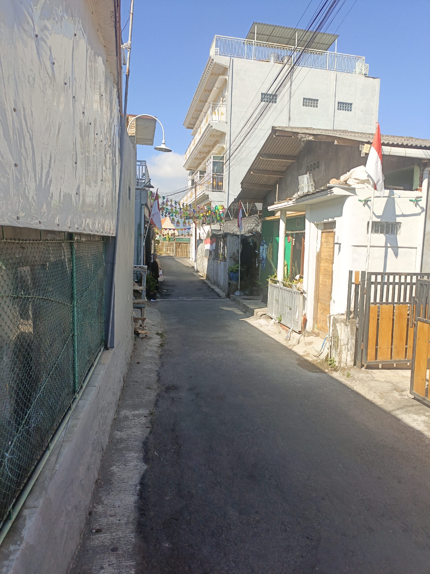 The image depicts a narrow asphalt road in the city of Batu, Indonesia, bordered by residential buildings and a fence. On the left, a large white tarp covers a wall, supported by a metal fence with green mesh. A street lamp with a curved white arm is mounted on a pole. On the right side of the road, a multi-story building with a light gray facade and balconies is visible, with a smaller building in front of it featuring a white wall and a wooden door. An Indonesian flag flies from a pole attached to the smaller building. Further down the road, colorful decorative banners are strung across the street, and a person in dark clothing is standing near the entrance of one of the buildings. The sky is clear and blue, indicating a sunny day. The scene appears to be in a residential area, possibly decorated for a local celebration.