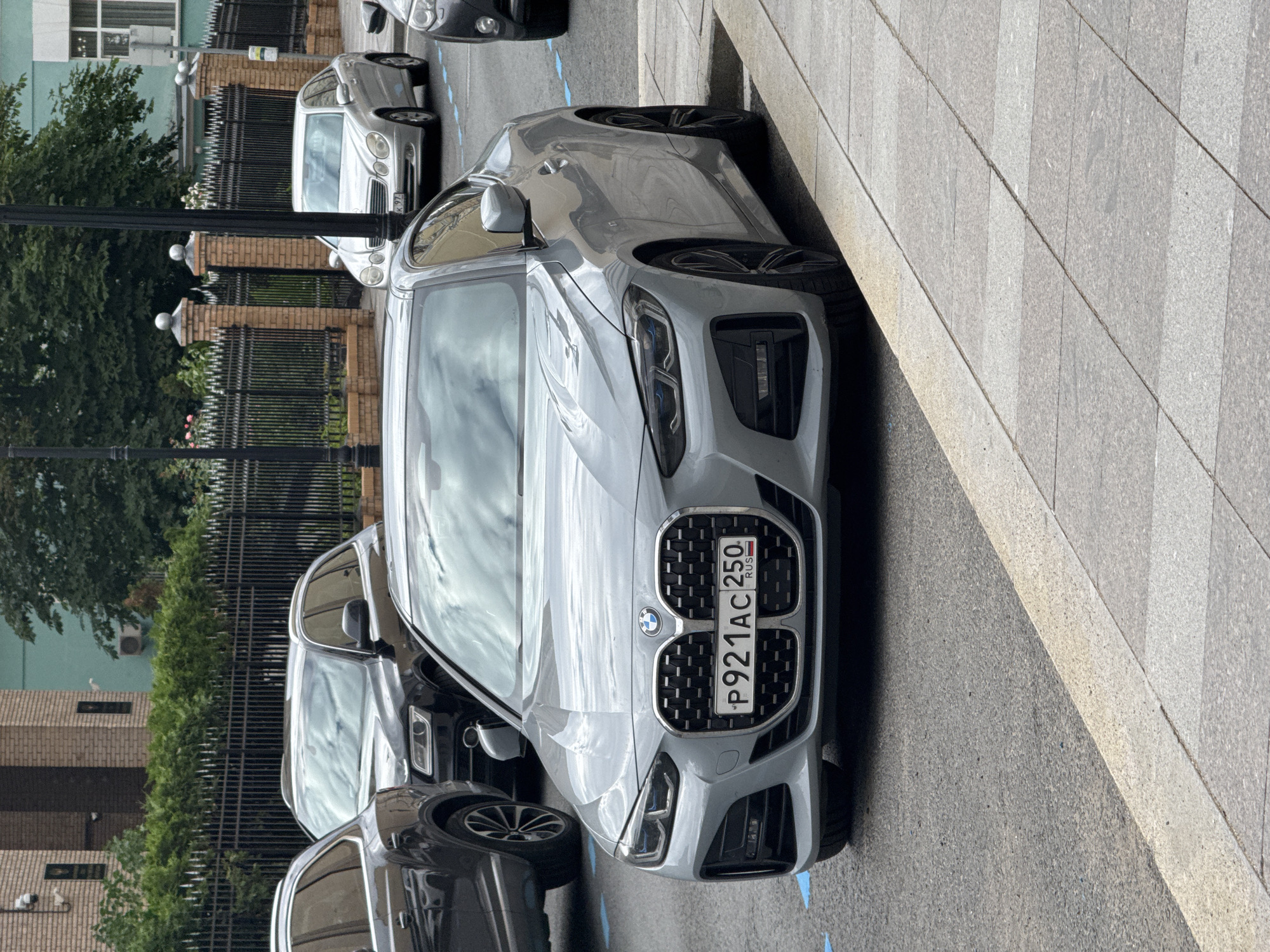 A silver BMW 4 Series coupe is parked on a street in Moscow, Russia. The car is positioned at an angle, with its front facing the camera. The license plate reads "P921AC 250 RUS". The street is paved, and a sidewalk with gray tiles is visible to the right of the car. In the background, a black metal fence with brick pillars separates the street from a green hedge and a building. Other cars, including a silver Mercedes-Benz sedan and a dark SUV, are parked further down the street. The sky is overcast.