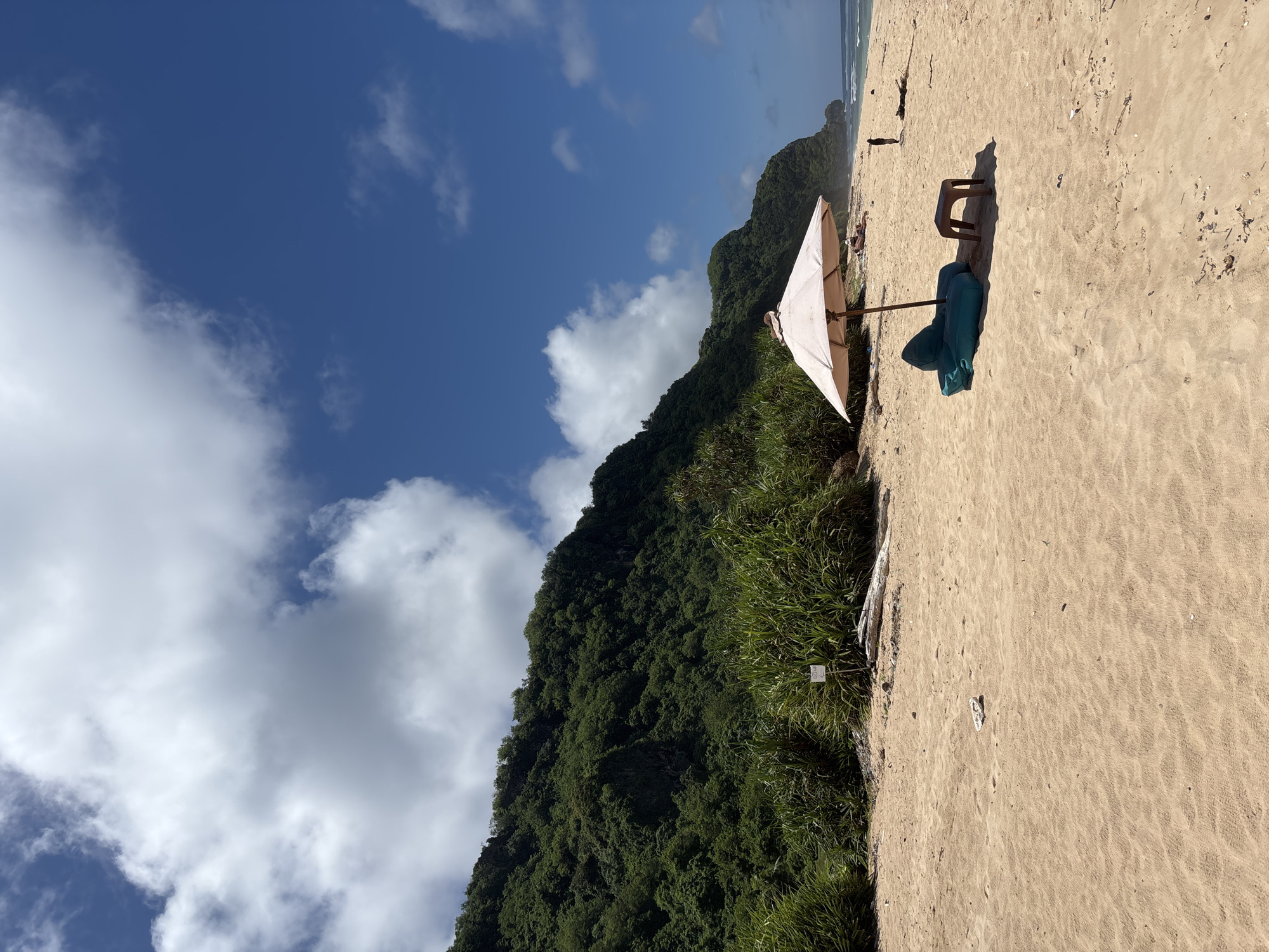 The image shows a beach in Pecatu, Indonesia, with a tan sandy shore. The sky is bright blue with scattered cumulus clouds. A mountain covered in green vegetation borders one side of the beach. An open white beach umbrella is situated near the edge of the vegetation. Next to the umbrella are a blue beach bag and a small brown stool. There is at least one person relaxing in the sand near the umbrella.