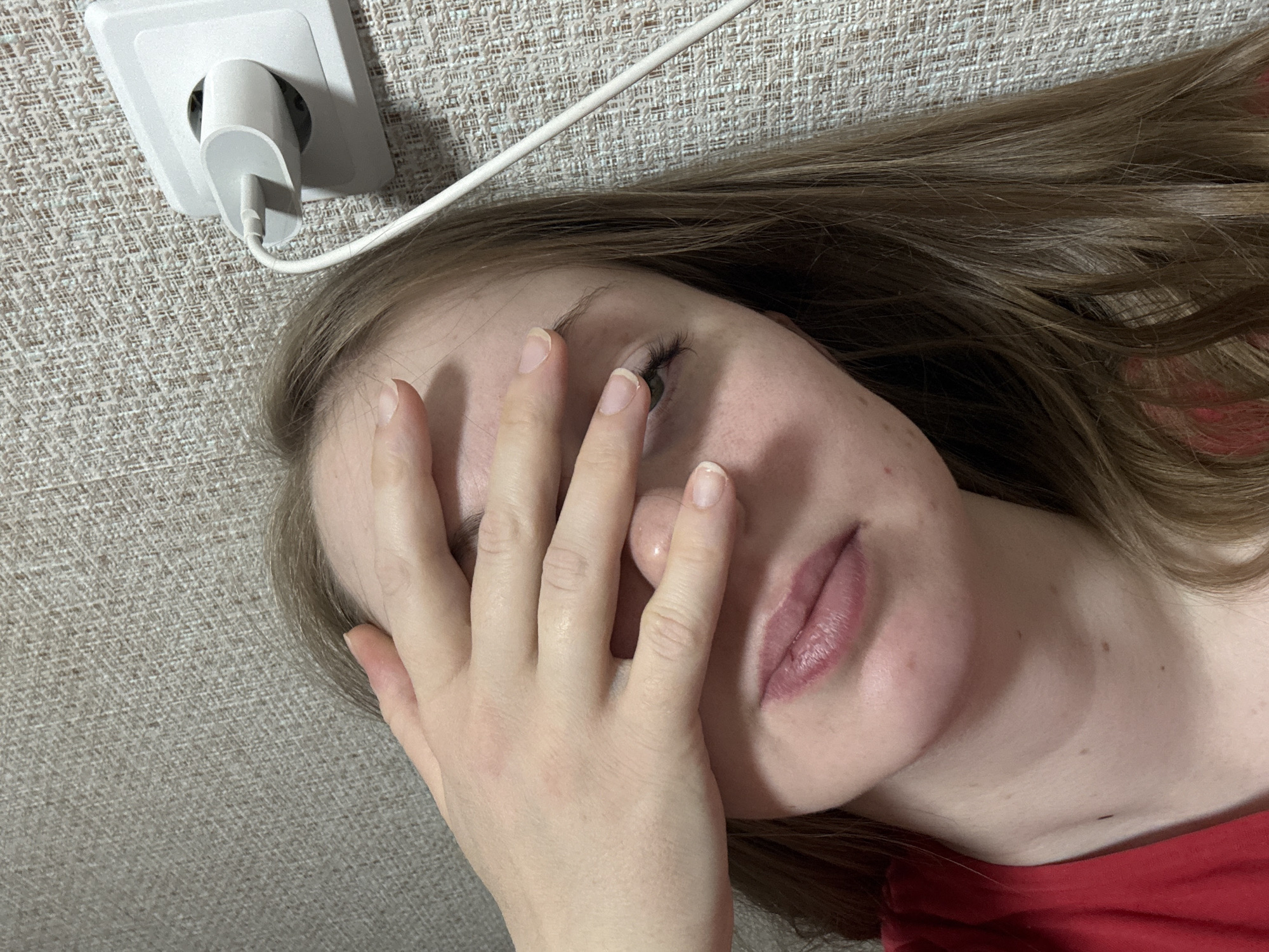 A fair-skinned female lies on a beige textured surface, possibly a couch or bed, with her right hand covering the upper half of her face. She has light brown hair that is spread out on the surface. A white electrical outlet and a charging cable are visible in the upper left corner of the image. The woman is wearing a red top. Location is Unknown, Unknown.