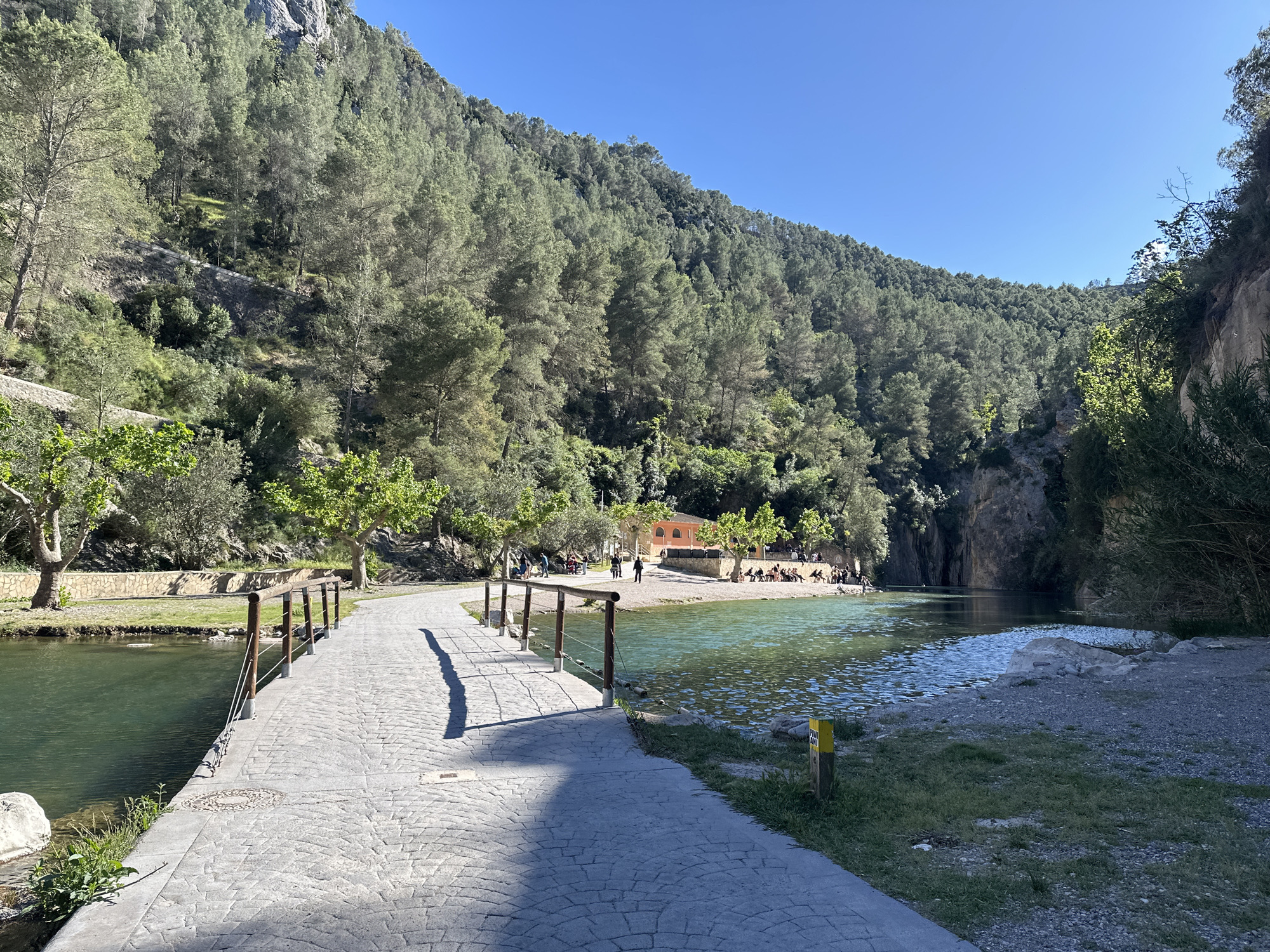 The image shows a scenic view of the Fuente de los Baños in Montanejos, Spain. A paved pathway extends from the lower left to the center of the frame, bordered by a wooden railing on one side and the edge of the water on the other. The pathway leads to a small beach area where people are present, some sitting, others standing or walking. A building with a peach-colored facade is visible near the beach. The water is calm and reflects the clear blue sky. The background is dominated by a steep, tree-covered hill that rises towards the left, with a cliff face visible on the right. The area is well-lit with sunlight.