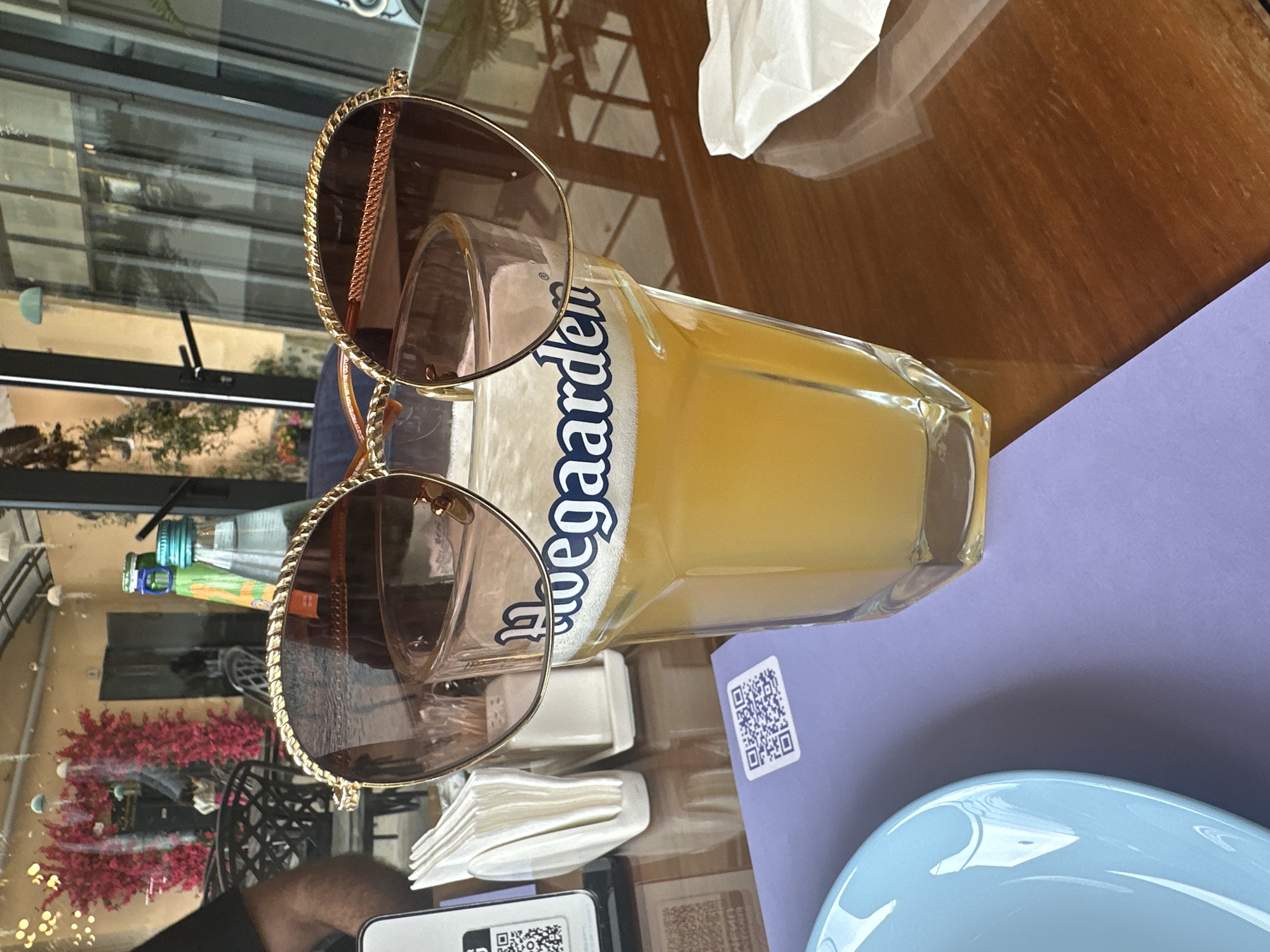 The image captures a scene on a table, likely in an outdoor dining area, with a distinctive blend of personal and culinary elements. A glass of what appears to be a wheat beer, identified by the label “Hoegaarden,” lies horizontally on the table. Perched on the glass is a pair of gold-rimmed sunglasses. The tabletop itself is a dark, polished wood. Adjacent to the beer glass, there's a stack of white napkins in a dispenser, a QR code on a purple placemat, and the edge of a light blue plate. In the background, a glimpse of what appears to be an outdoor setting is visible through a window or open doorway. This setting seems to be near an outdoor garden, possibly a restaurant's patio, adorned with bright red flowers. The reflection in the sunglasses suggests an urban environment with buildings, possibly located in Rekha, Georgia. In the bottom left corner, a person in a dark shirt, whose arm is visible, is seated at the table, holding a smartphone with a partially visible QR code on the screen.