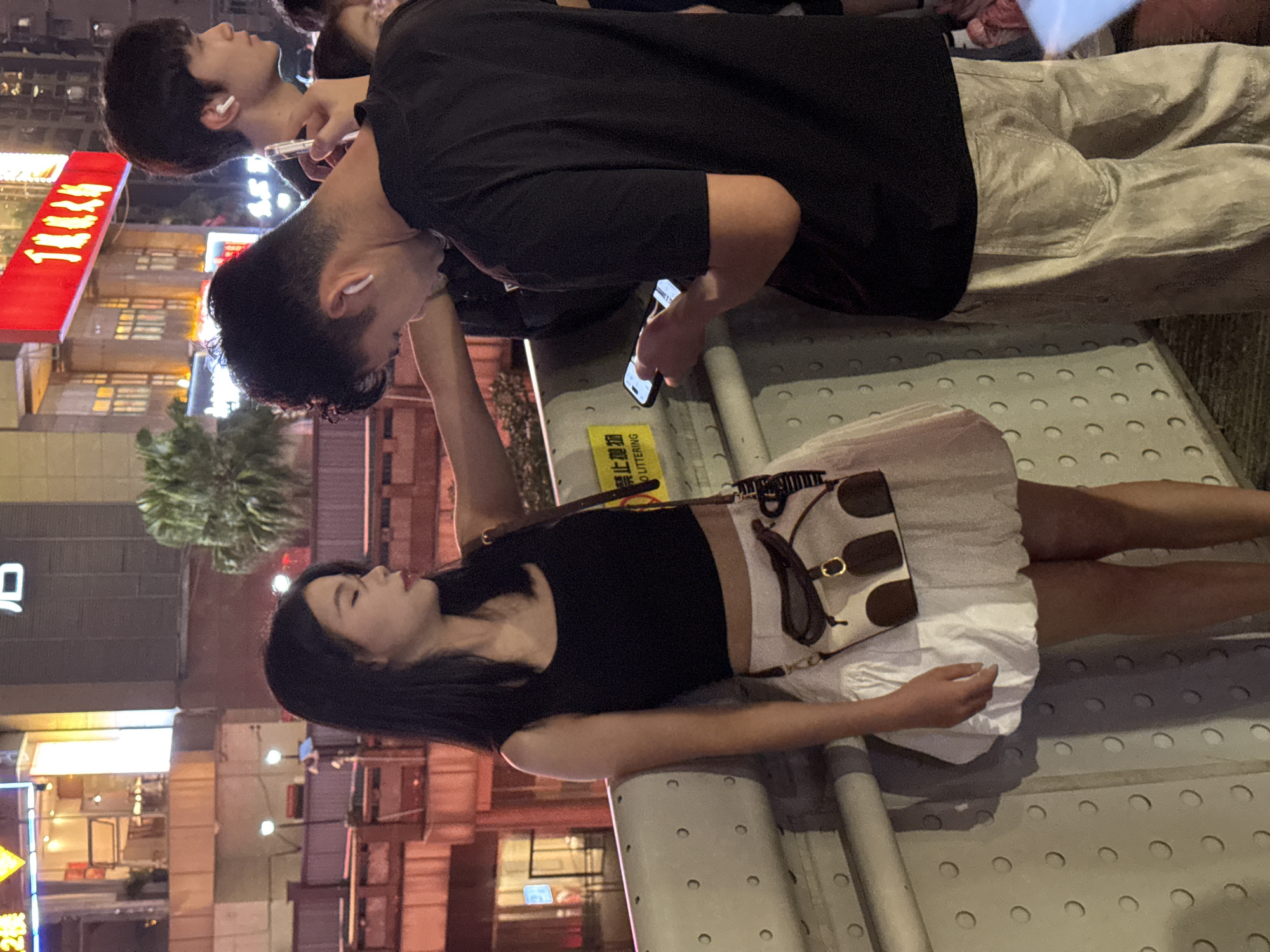 Two young adults are standing on a bridge or elevated walkway in Yuzhong District, China, at night. The woman on the left, with long dark hair, wears a black crop top and a white ruffled skirt. She has a small crossbody bag with brown accents. The man on the right, also with dark hair, wears a black t-shirt and light-colored pants. He is holding a smartphone and appears to be interacting with it. Both individuals have white wireless earbuds in their ears. In the background, there are illuminated buildings and a prominent red neon sign with Chinese characters. The walkway has a white railing with circular indentations. A yellow sign with Chinese text and the English words "NO LITTERING" is visible. The lighting suggests an urban environment with artificial illumination from the buildings and signs.