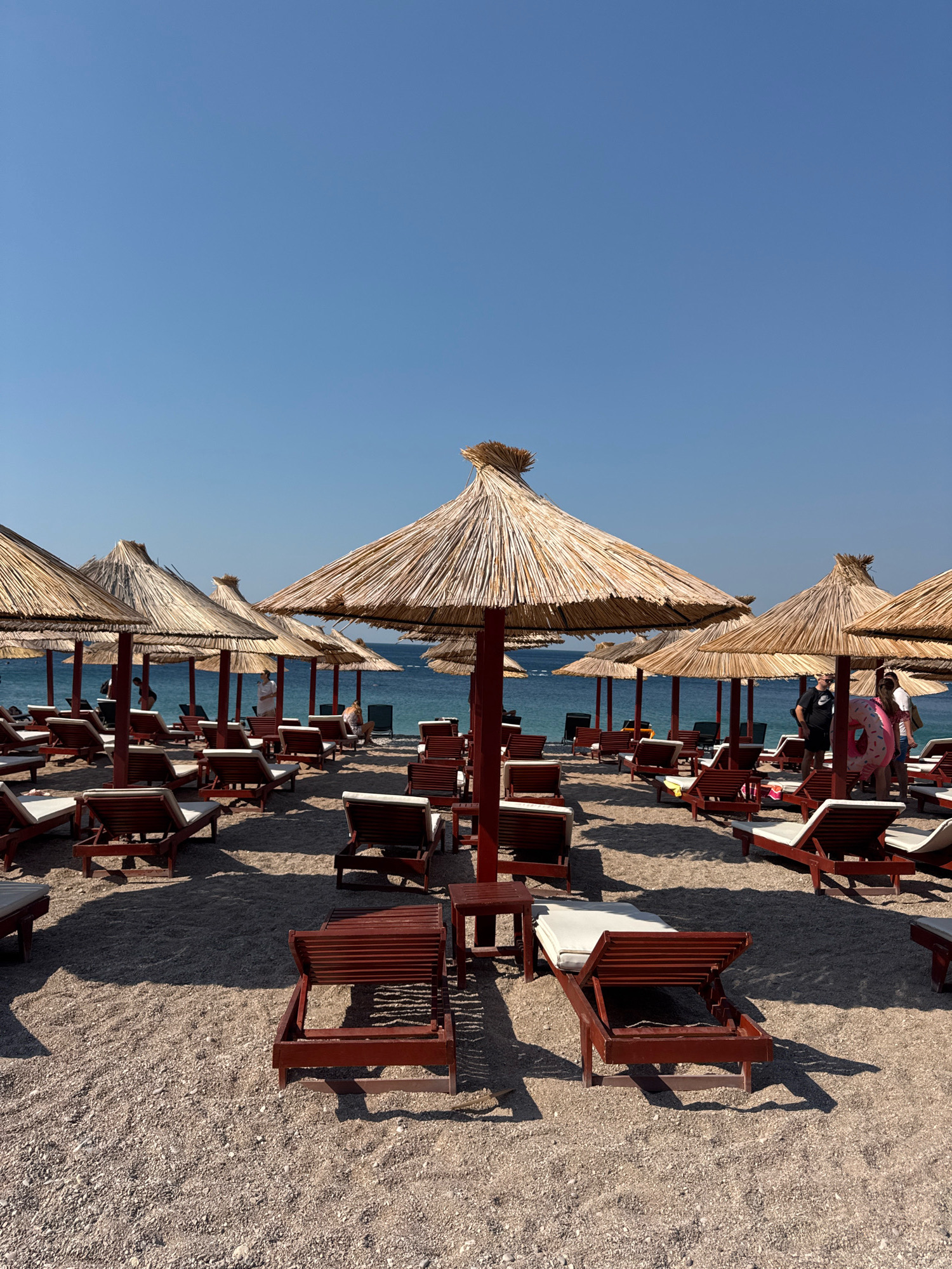 The image depicts a beach scene in Mirište, Montenegro, characterized by numerous empty lounge chairs arranged on a pebbly shore. Several thatched umbrellas provide shade over the seating areas. In the background, the calm blue sea meets a clear sky, with a few distant people visible near the water's edge. The overall atmosphere is serene and typical of a beach resort.