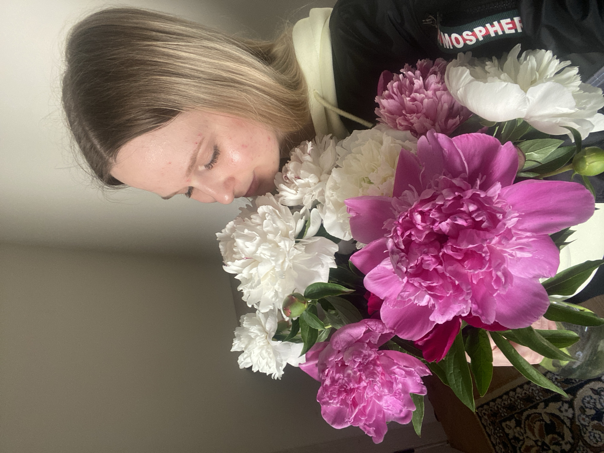 The image shows a close-up, eye-level indoor portrait of a female engaging with a large bouquet of peonies. The subject, a woman with light skin and shoulder-length blonde hair exhibiting darker roots, has her eyes closed and head tilted, bringing her nose into direct proximity with the flowers, indicating the action of smelling them. Minor facial blemishes are visible on her forehead and cheeks. She is wearing a dark-colored jacket or top, under which a light yellow or cream-colored hood is visible. On the right sleeve of her jacket, a decorative band with green, white, and red lettering is present, partially spelling "MOSPHF".
The bouquet comprises a mixture of fully bloomed and partially open peonies, featuring both vibrant pink/magenta and white varieties, interspersed with green leaves. A single green flower bud is also visible among the blooms. The lower section of a clear glass vase containing the flowers is partially visible at the bottom-center of the frame. The background consists of a plain, light-toned wall, while a segment of a patterned dark rug is visible on the floor in the lower-left corner of the image. The scene is illuminated by what appears to be a natural light source, casting soft shadows. The location is Unknown, Unknown.