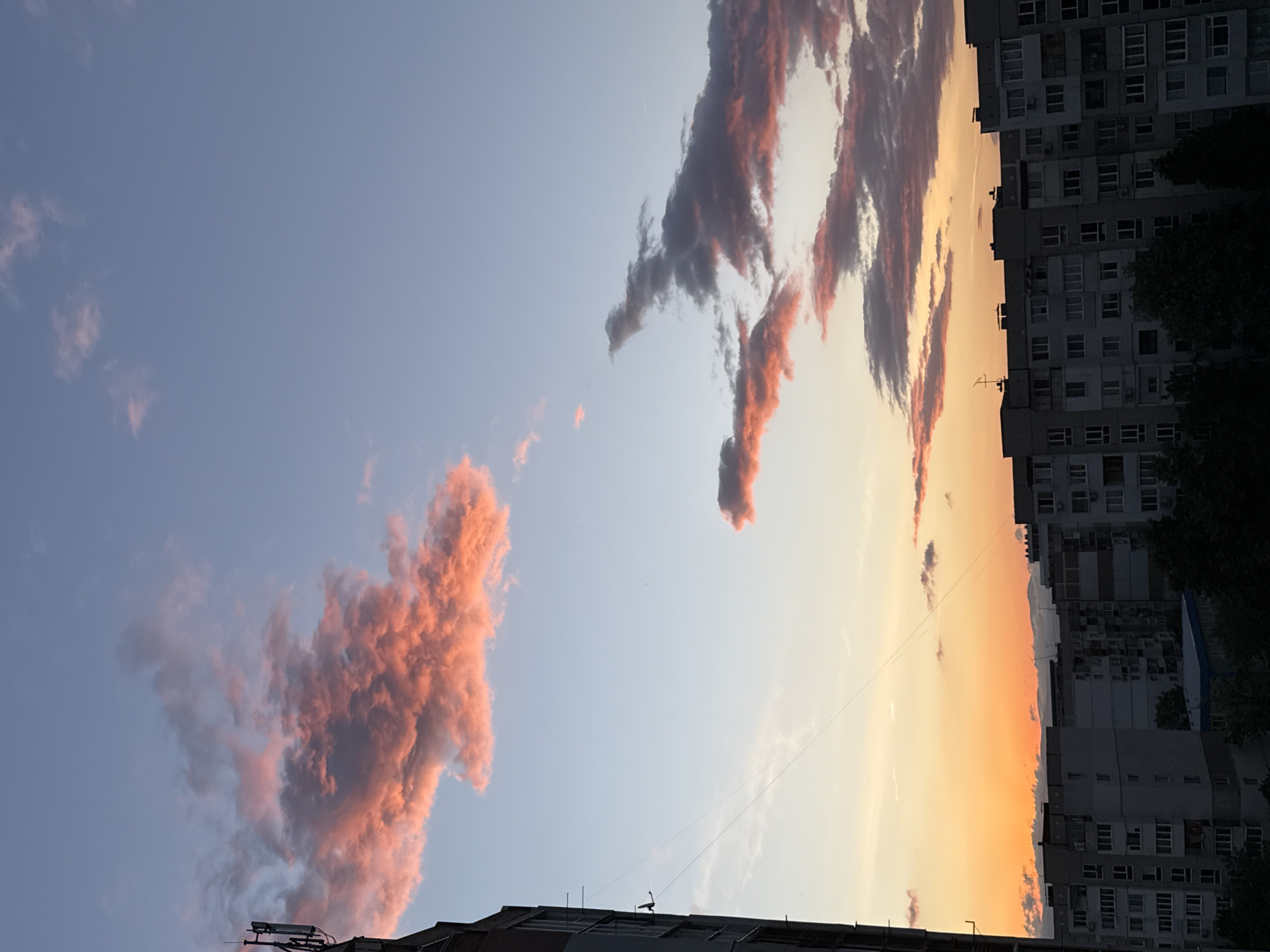 The image shows a view of the sky at sunset in Sofia, Bulgaria. The sky is a mix of blue and orange, with pink and grey clouds scattered across it. Buildings are visible in the foreground and right side of the image. The buildings appear to be residential apartment blocks.