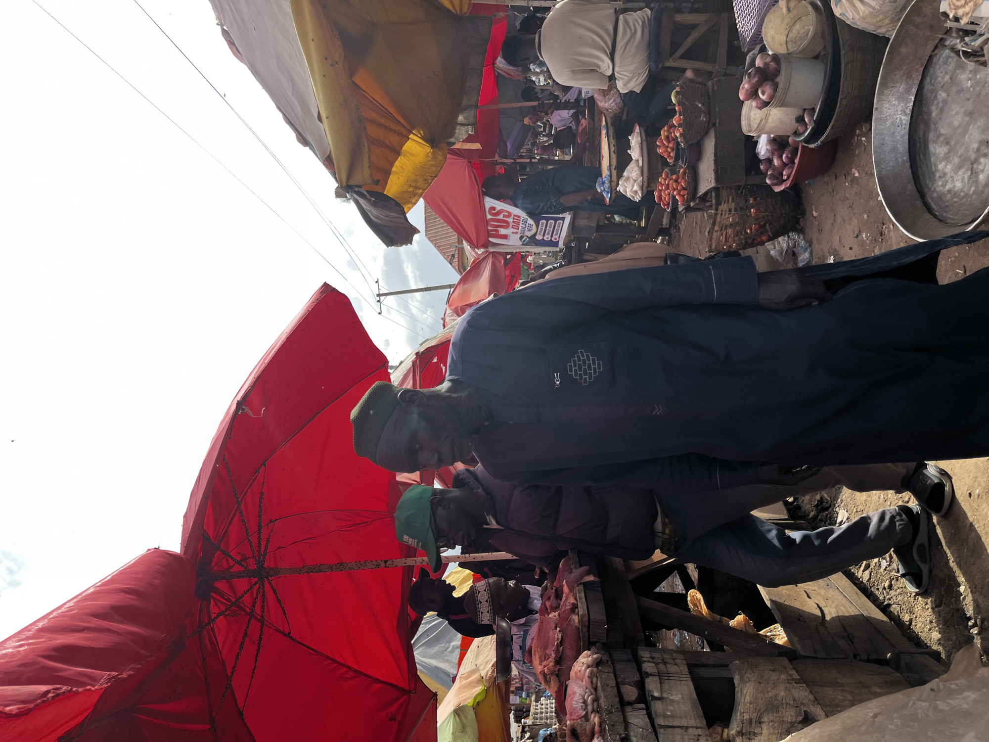 The image depicts a bustling market scene, likely in Rafin Par, Nigeria, with several individuals engaged in commerce. In the foreground, a man wearing a dark blue, long-sleeved garment and a dark cap walks towards the viewer, his gaze directed downwards. To his left, another man in a dark jacket and a green baseball cap with the word "YOU" visible stands near a stall laden with raw meat. He is looking towards the meat, possibly a vendor or customer. Behind them, other marketgoers and stalls are visible, indicated by the presence of red and yellow umbrellas providing shade. The ground is dirt and appears somewhat uneven, with various goods, including baskets of tomatoes and onions, displayed. The overall atmosphere suggests a lively outdoor marketplace.