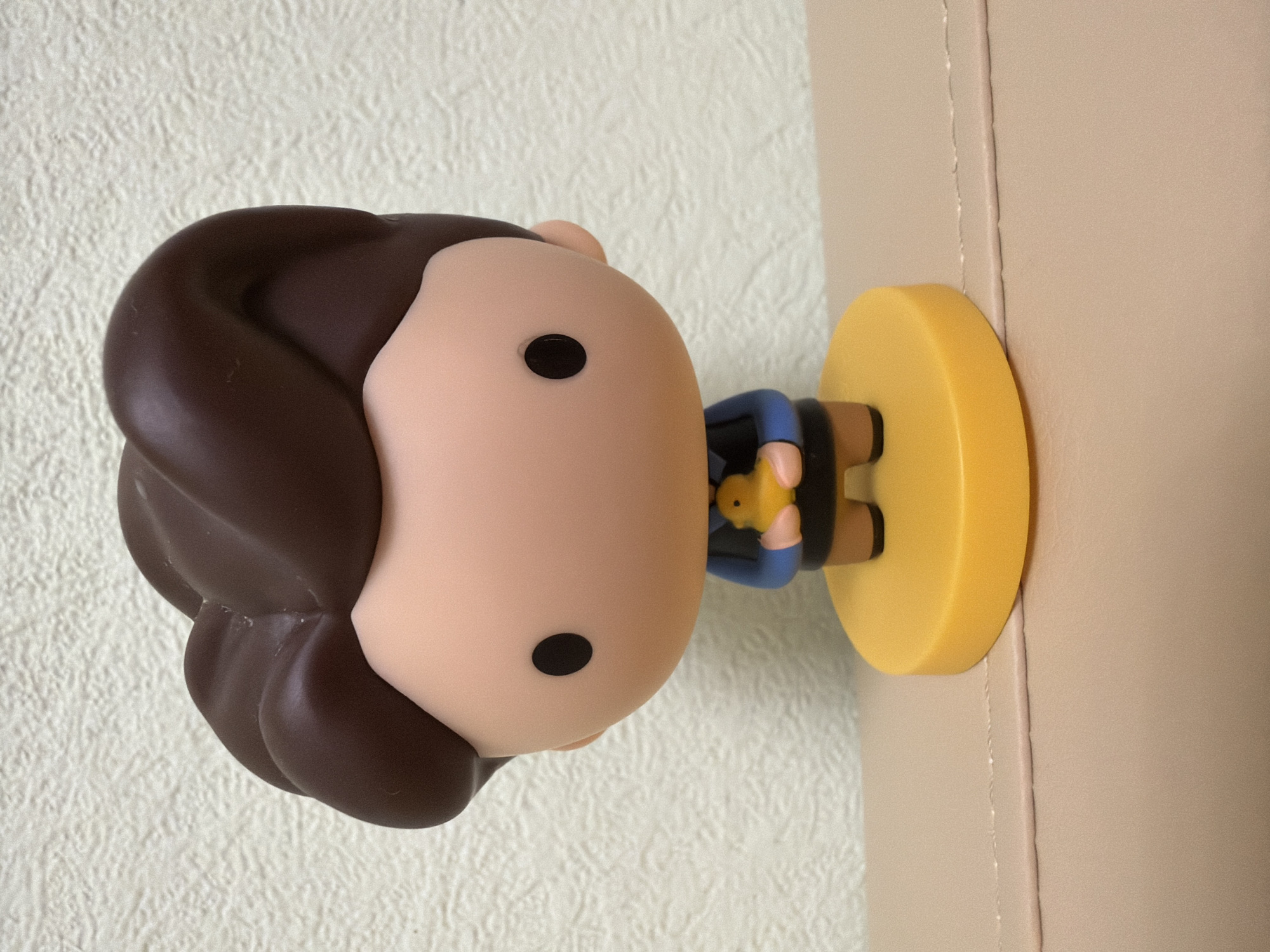A small, cartoonish figurine with brown hair, a light brown face, and black eyes stands on a bright yellow disc base. The figurine is wearing a dark blue jacket with a black collar and holds a small yellow duck in its hands. The figurine is positioned in the center of the frame. The background consists of a textured, light-colored wall. The figurine rests on a smooth, light brown surface.