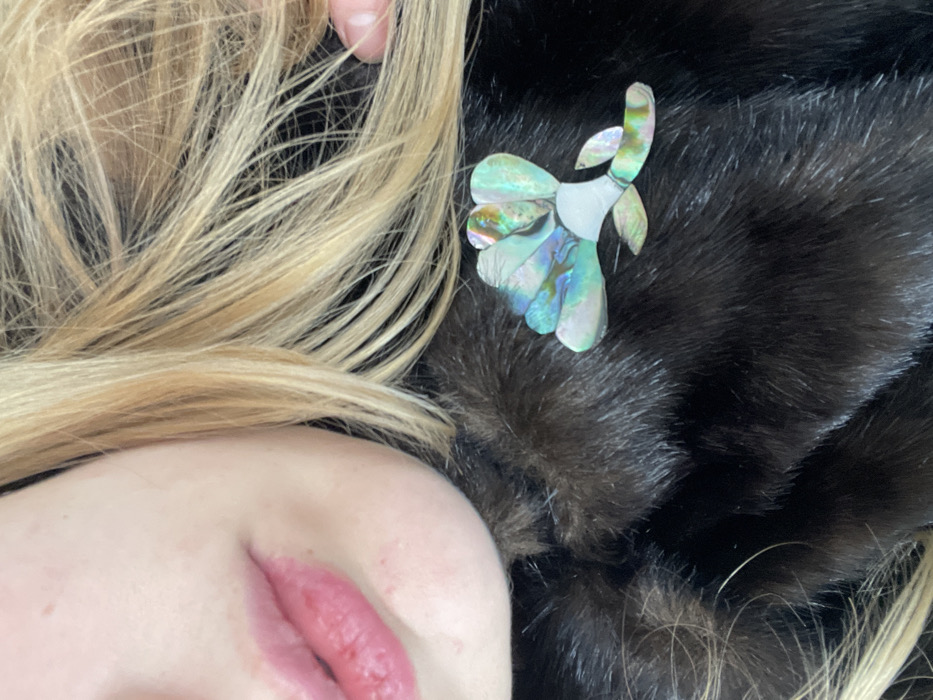 A close-up photograph captures the lower face, right shoulder, and upper chest of an individual. The subject exhibits fair skin and blonde hair, which partially obscures the dark, plush fur or faux fur garment worn. The lips are reddish, appearing somewhat chapped. Pinned to the fur garment, on the right side of the frame, is a stylized flower-shaped brooch. This brooch is composed of multiple iridescent pieces, likely abalone shell or mother-of-pearl, displaying hues of green, blue, purple, and brown, along with opaque white segments forming its central structure. A fingertip is partially visible in the upper right quadrant of the image, near the blonde hair. The background is indistinct and offers no environmental context. The specific location within London, United Kingdom, cannot be ascertained from the visual evidence provided.