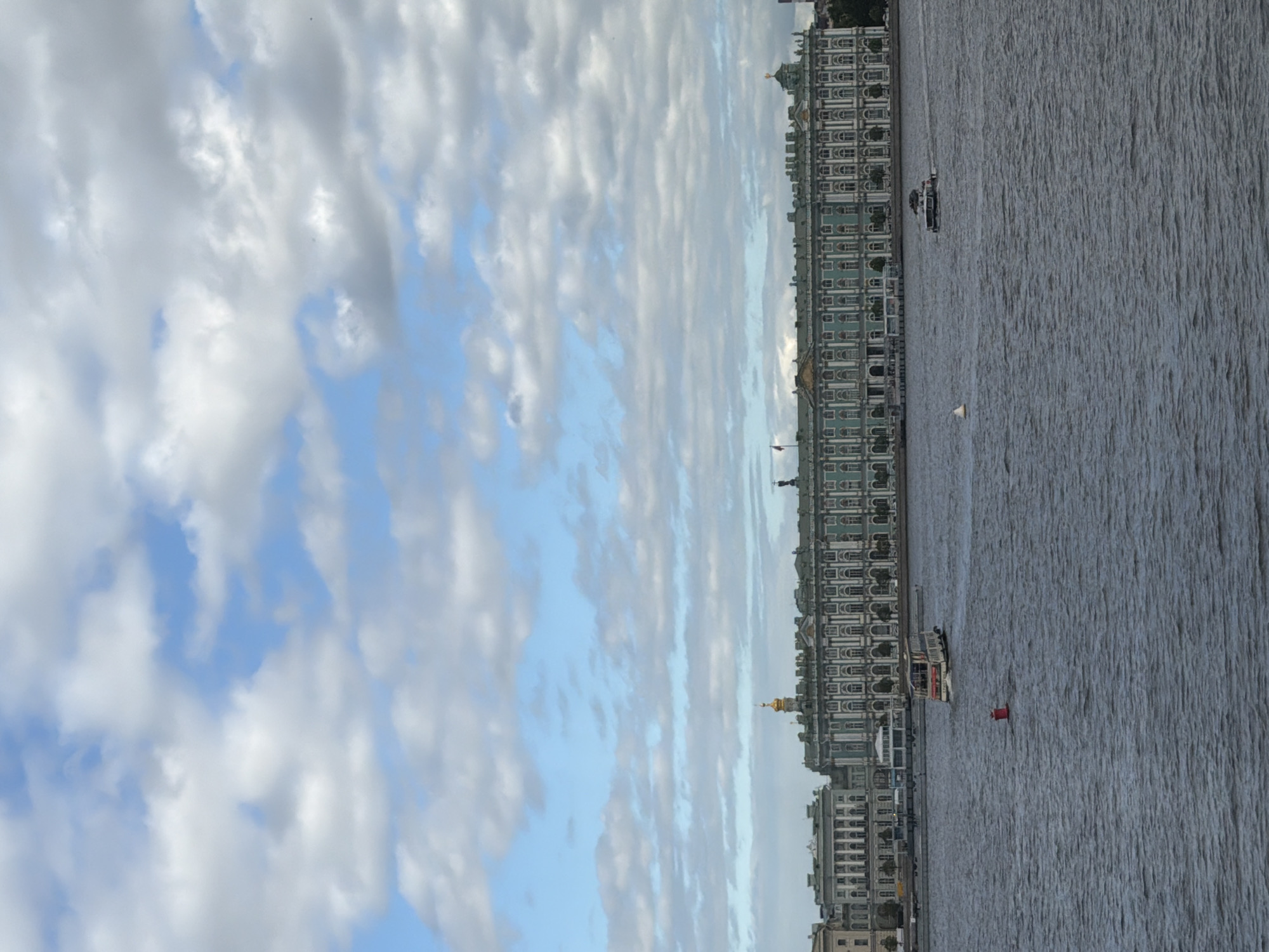 This media file displays a scene featuring a body of water, a large building complex, and a cloudy sky. The building appears to be an ornate palace or government structure with many windows and a green facade, stretching along the edge of the water. There are several boats on the water, including a larger passenger boat with people visible on its deck, and a smaller vessel. A red buoy is also present in the water. The sky is filled with dramatic, textured clouds in shades of white and gray, with patches of blue sky showing through. The lighting suggests it might be late afternoon or early evening, or a generally overcast day. The orientation of the image is landscape. The overall impression is one of urban scenery with a waterfront setting.
