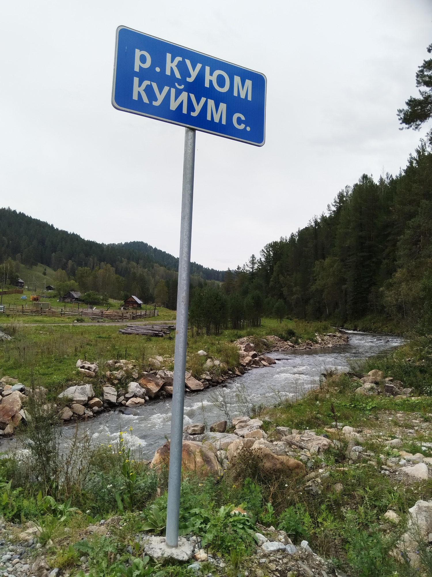 A blue sign with white Cyrillic text is mounted on a metal pole in the foreground. The text on the sign reads "р. КУЮМ КУЙУМ с.". The sign is positioned to the left of a rural landscape that includes a river, trees, hills, and some wooden structures. The river is flowing from right to left in the image, with rocky banks and some vegetation. In the mid-ground, there is a green field with several wooden buildings and fences, resembling a small farm or village. Behind the field, there are forested hills that extend to the horizon under a cloudy sky. The overall impression is one of a remote, natural setting in a rural area. The location is identified as Nizhnii Kuium, Russia, based on the sign text. There are no people visible in the image.