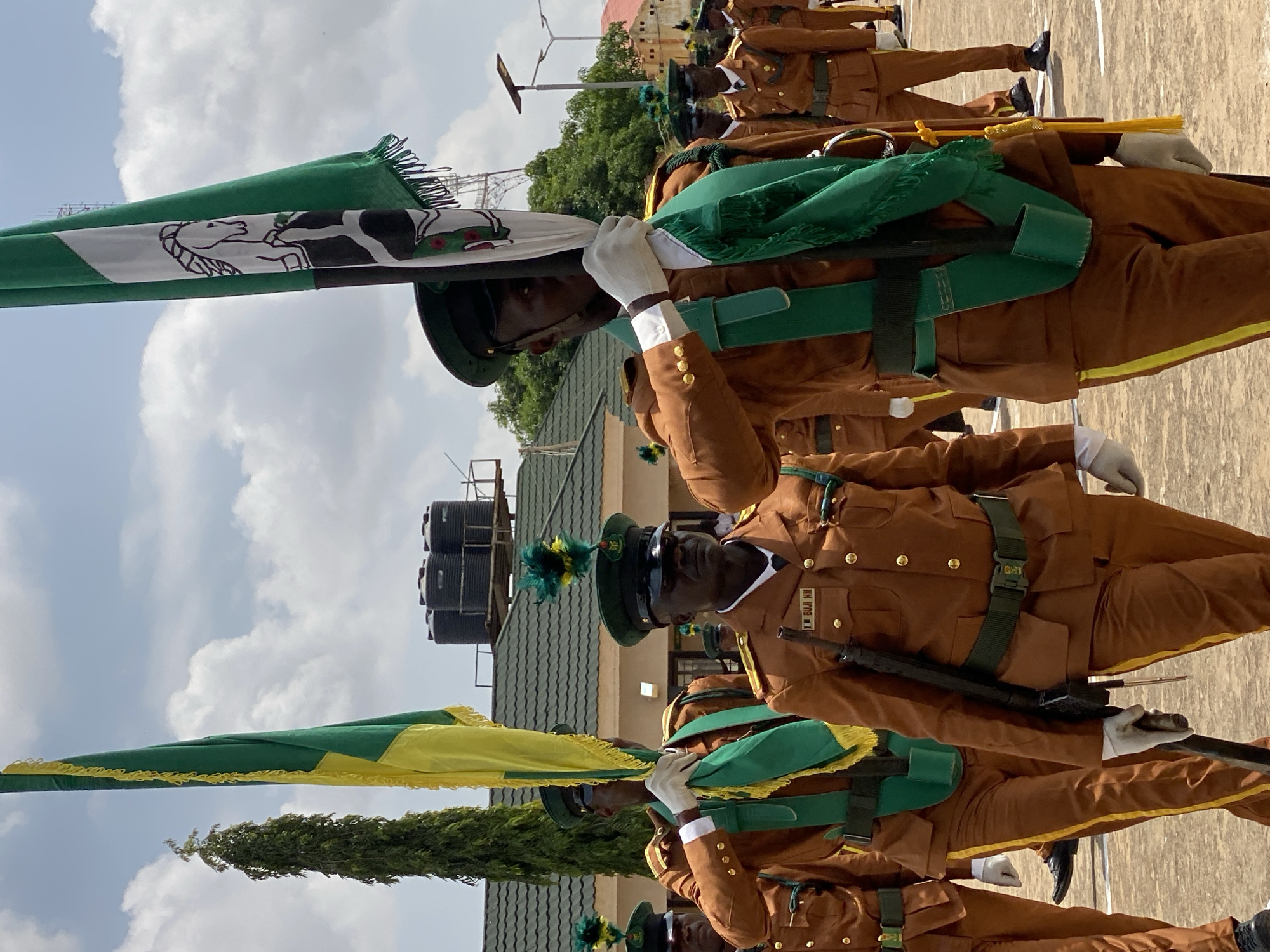 This is a vertical, medium-shot photograph taken outdoors on a cloudy day, likely during daylight hours. The scene depicts a group of men in uniform, participating in a formal parade. They are dressed in brown uniforms with green sashes and dark green caps adorned with colorful feathers. Some of the men are holding flags. One flag is green and white with an illustration of a horse and a cow. Another flag is green and yellow. The men are wearing white gloves and appear to be carrying rifles.
The background shows a building with a green, tiled roof, and a clear sky with scattered clouds. There are also large water tanks visible in the background, suggesting an institutional setting. The overall atmosphere seems serious and orderly, befitting a military or ceremonial event. The image captures a moment of marching or standing at attention during the parade. There is visible text on a nametag worn by one of the individuals, which reads "I BUI NM". The location context is Kaduna, Nigeria.