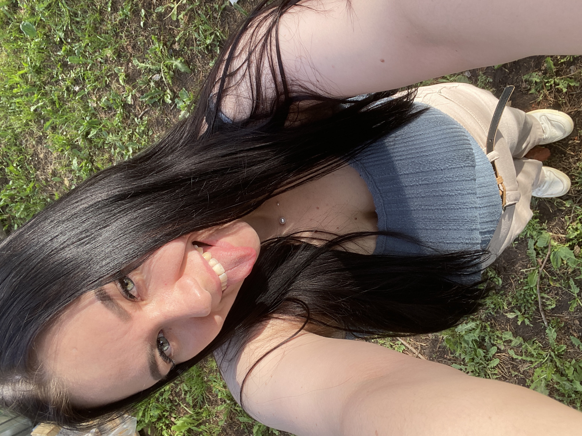 A young adult woman is posing for a selfie outdoors. She is lying on the ground, likely grass, as indicated by the green vegetation in the background. She is wearing a light blue knitted top and beige pants with a belt. Her hair is long, black, and shiny. She is sticking out her tongue in a playful manner. She is wearing white sneakers. The location is the city of Chaikovsky, Russia.