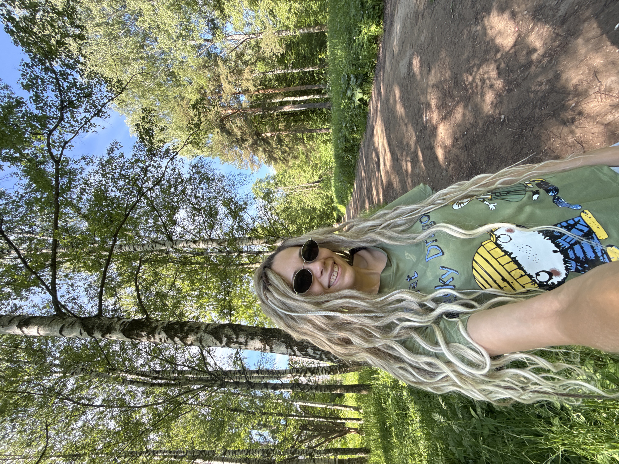 An outdoor, eye-level, medium-shot photograph captures a woman with long, blonde, braided hair and sunglasses smiling while holding the camera. She is standing in a park-like setting with birch trees and a dirt path in the background. The woman is wearing a green t-shirt with a cartoon character on it. The sun is shining, casting shadows on the ground and illuminating the lush greenery. The location is identified as Rusanovo, Russia.