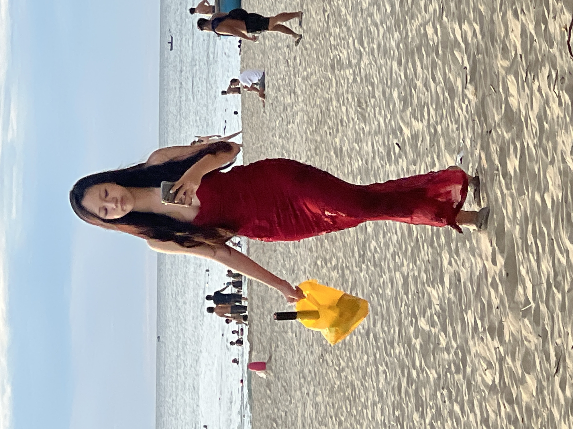 A young woman in a long, fitted, red dress walks on a sandy beach. She is holding a yellow plastic bag in her left hand and a mobile phone in her right hand, looking down at the phone. The beach is populated with other beachgoers in the background, some in the water, and others sitting or standing on the sand. The ocean is visible behind them under a bright sky with scattered clouds. The scene is likely located on one of the popular beaches of Đà Nẵng, Vietnam, such as My Khe Beach, known for its wide sandy shores and vibrant atmosphere.