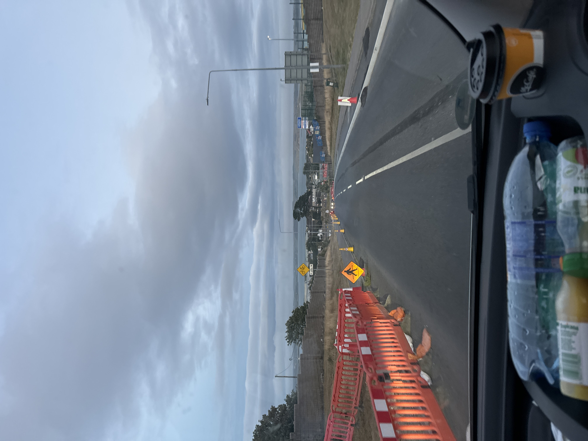 The image is taken from the interior of a vehicle, looking forward along a road that appears to be undergoing construction or maintenance. In the foreground, on the dashboard, are two plastic water bottles and a McCafe coffee cup with a straw. The road ahead is asphalt with white lane markings. To the left of the road, there is a construction barrier made of orange plastic fencing with white reflective strips, illuminated by orange lights. Several yellow traffic cones and warning signs are visible along the roadside, including a sign depicting a pedestrian and a sign with a bicycle symbol. In the distance, a body of water is visible, with land and buildings on the opposite shore. The sky is overcast with heavy clouds.
Based on the presence of the McCafe cup and the general landscape, the location is likely in or around Tramore, Ireland, a coastal town known for its beaches and seaside attractions. The construction and the road suggest a public thoroughfare.
