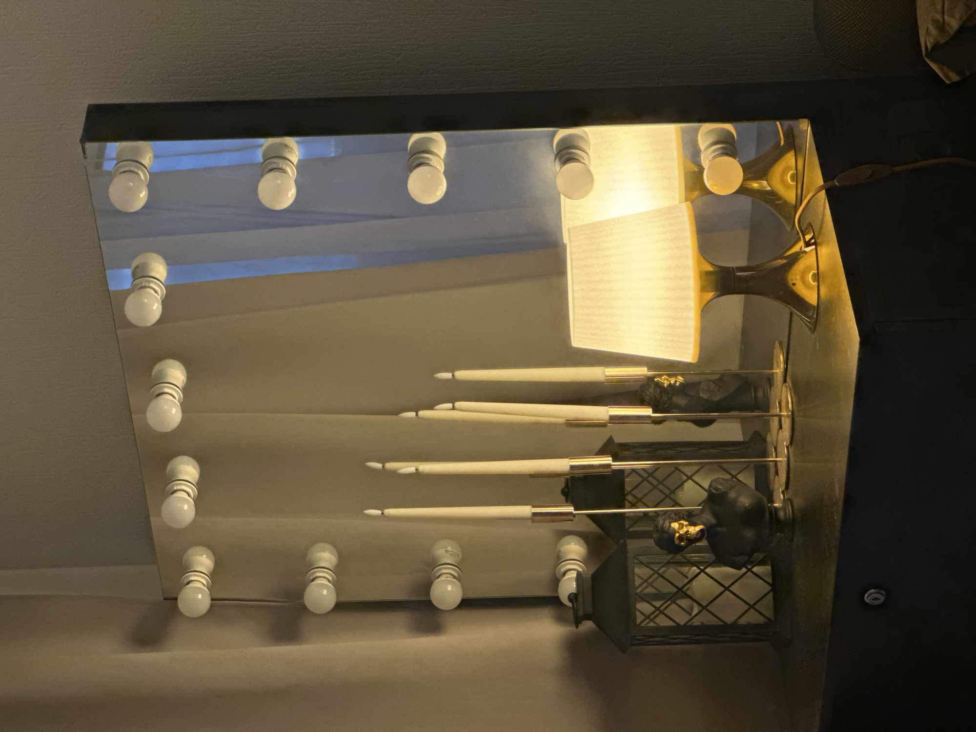 The image depicts a mirror reflecting a room interior. The mirror has a row of light bulbs around its perimeter. The reflection shows a lamp with a lampshade and a decorative object with candles. The scene suggests an indoor setting, possibly a dressing room or vanity area. The location is not discernible from the image.